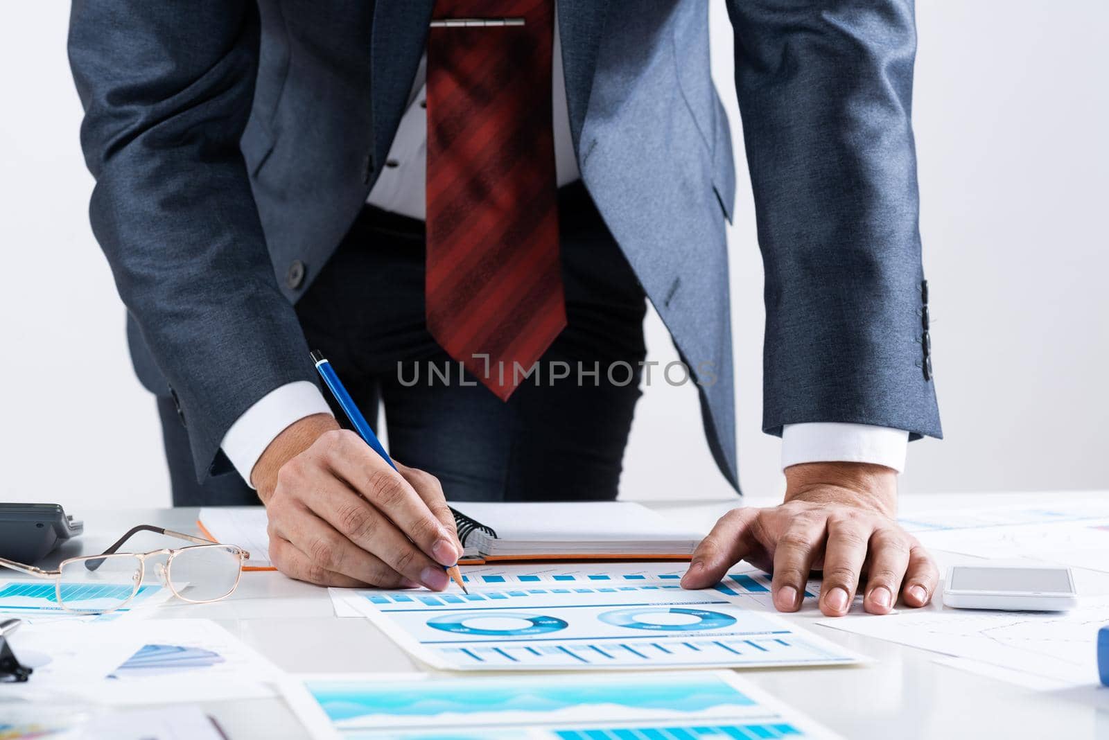 Businessman standing near office desk and analyzing financial charts. Successful corporate manager in business suit and tie. Close-up man hand with pen. Entrepreneurship and leadership concept.