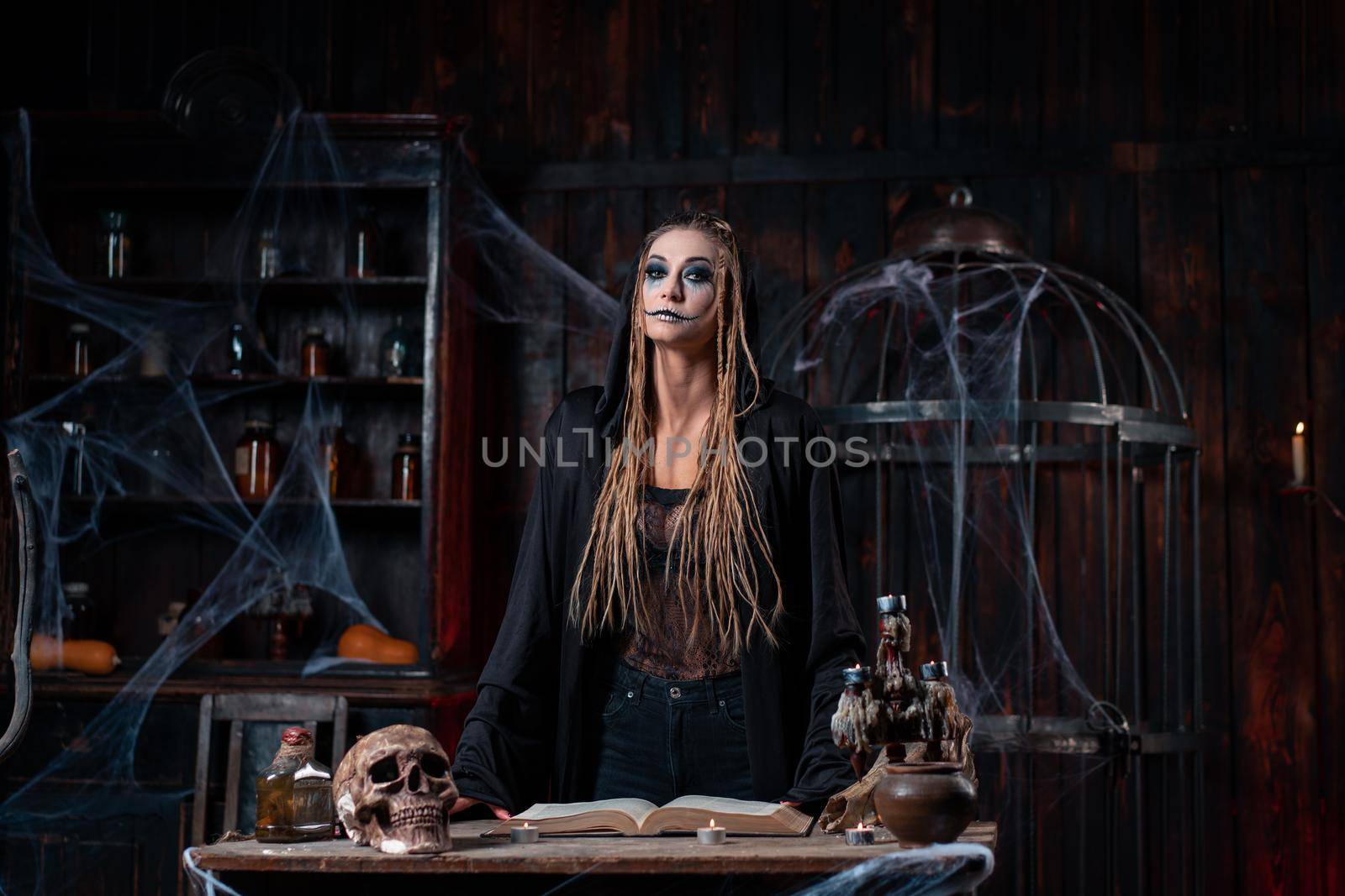 Halloween concept. Witch dressed black hood with dreadlocks standing dark dungeon room use magic book for conjuring magic spell. Female necromancer wizard gothic interior with skull, cage, spider web