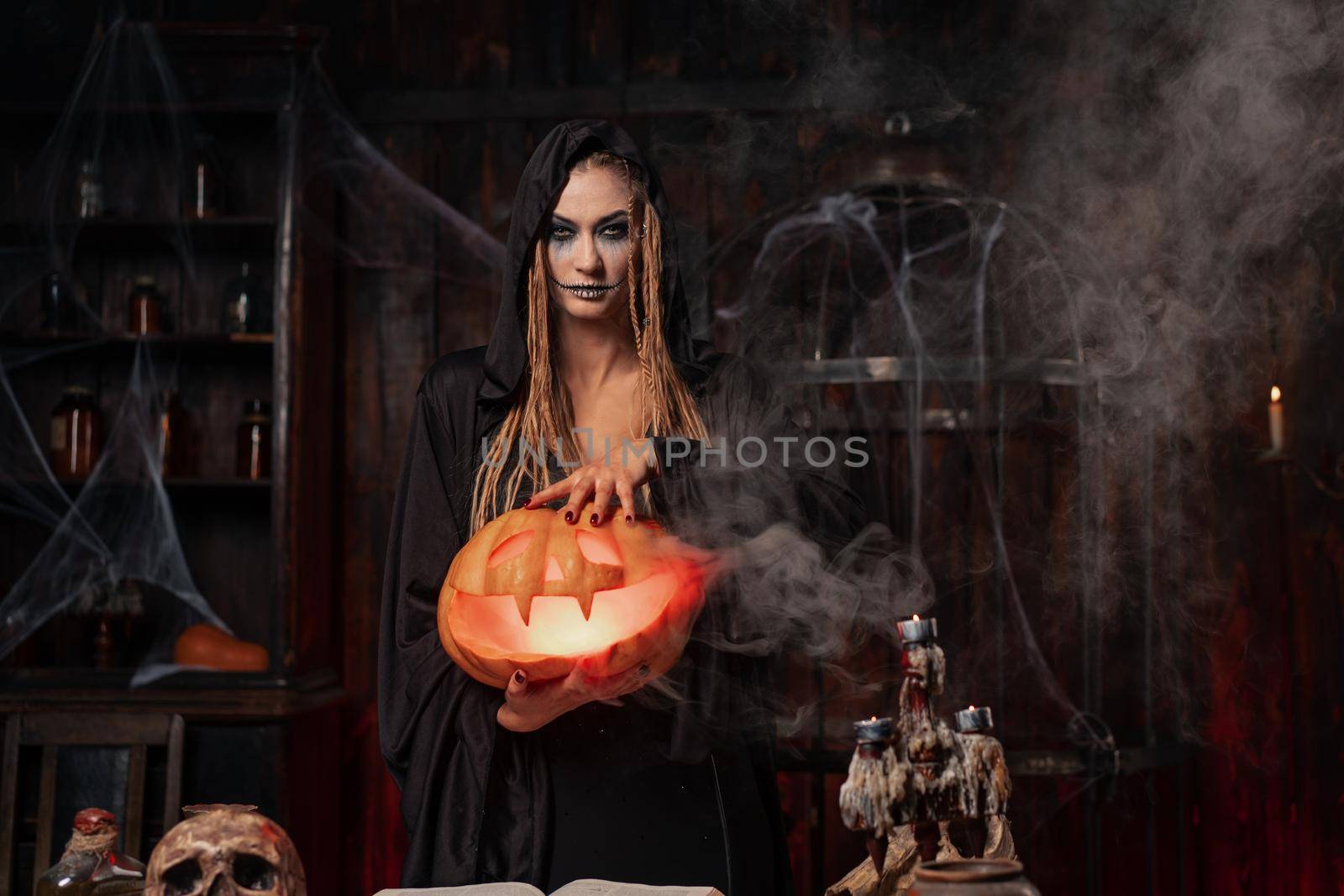 Halloween concept. Black witch holding Halloween pumpkin in hand with carved smily face in hand standing dark room. Female necromancer, dungeon place. Jack o lantern head.