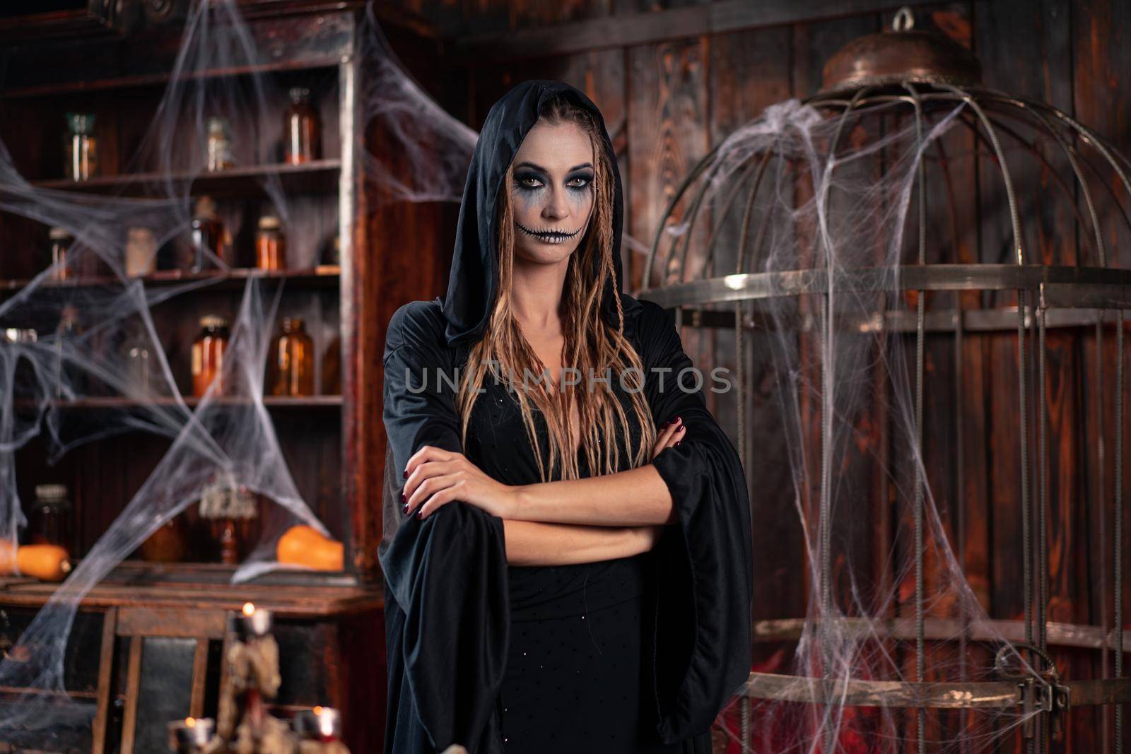 Halloween concept. Witch dressed black hood with dreadlocks standing dark dungeon room conjuring magic spell. Female necromancer wizard gothic interior with skull, cage, spider web