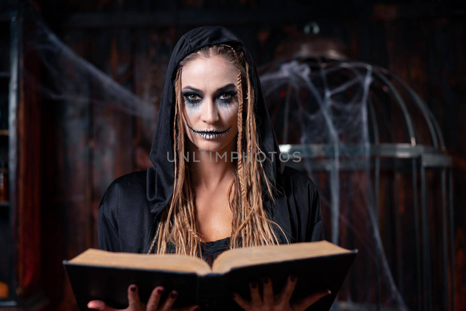 Halloween concept. Witch dressed black hood with dreadlocks standing dark dungeon room use magic book for conjuring magic spell. Female necromancer wizard gothic interior looking camera close up