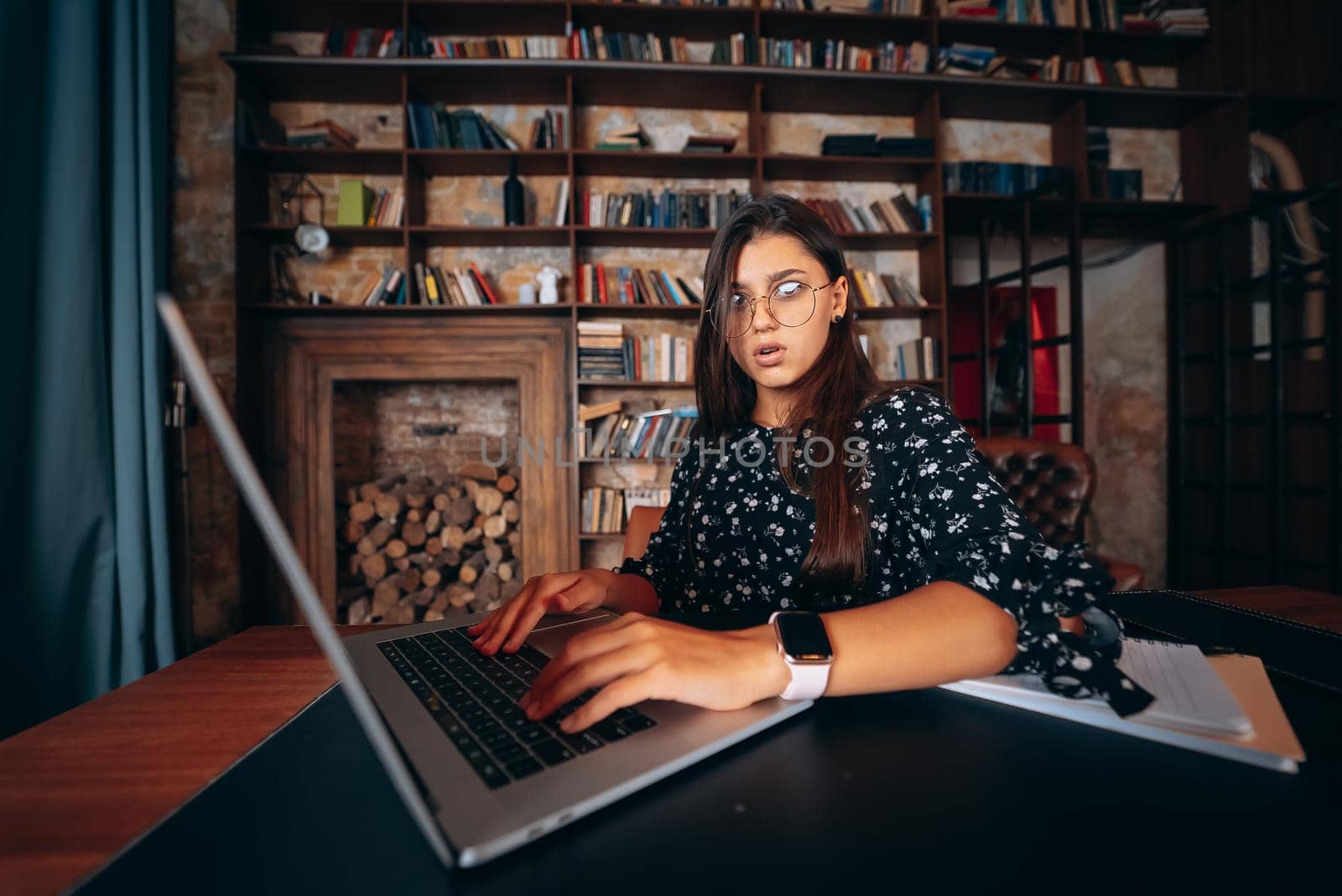 Young beautiful woman in glasses works at the laptop while sitting at the table