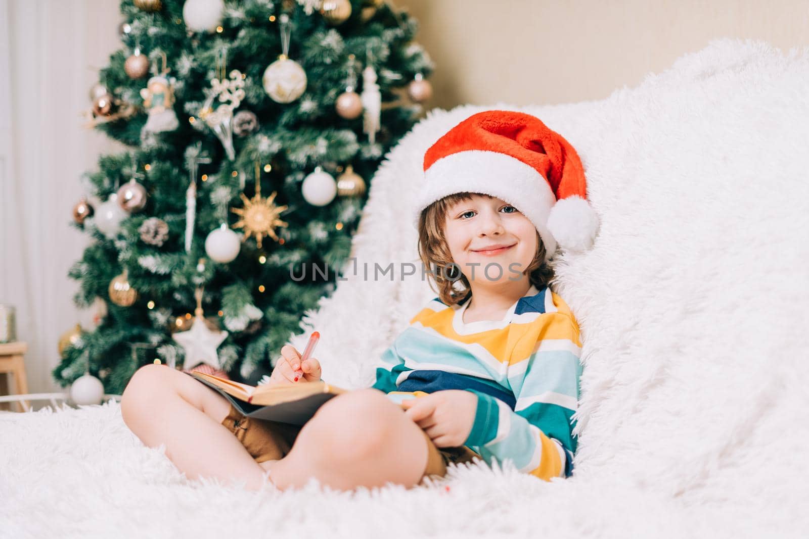 Smiling Kid boy in red Santa hat sit on couch and writing the letter to dear Santa at home. Child wish list. Dreams of a Christmas gifts. Merry Christmas and Happy new year