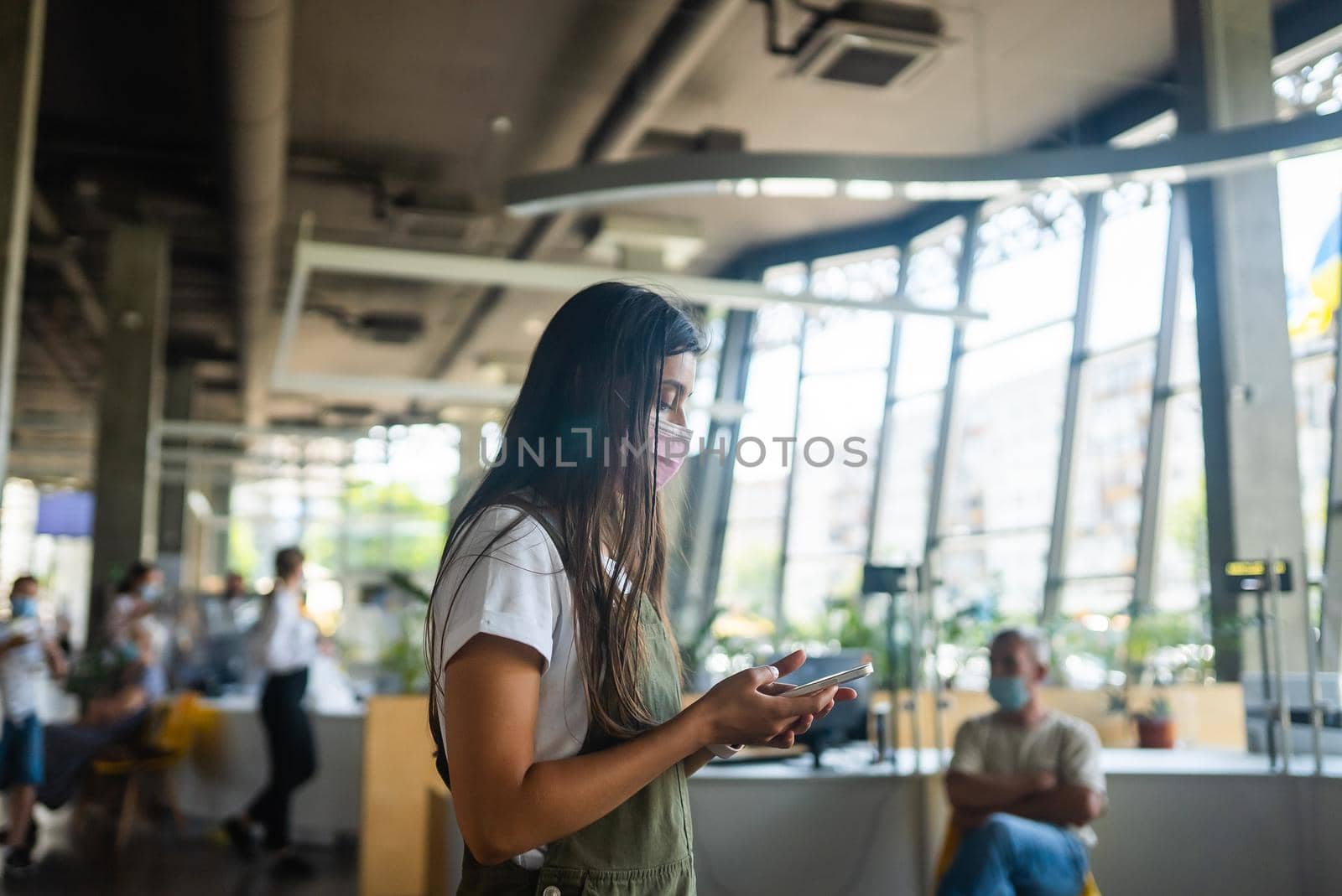 Portrait brunette girl in pink protective medical face mask uses smartphone at indoor.