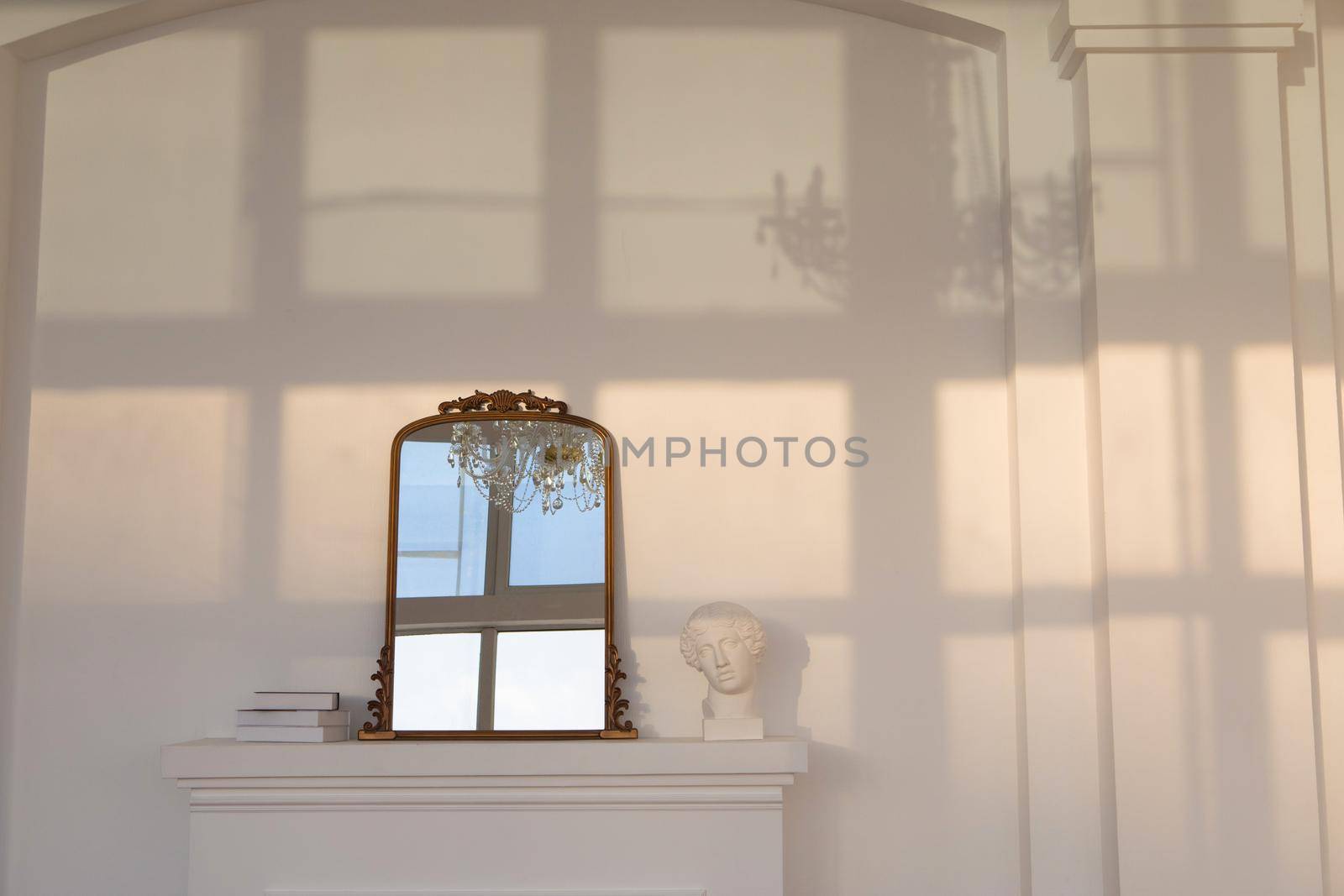 Mirror placed on artificial white fireplace near statuette and stack of books against white wall with shadow of window and chandelier on it
