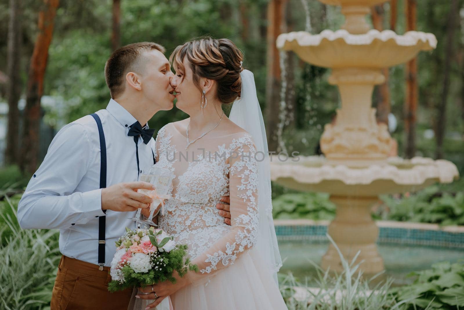 bride and groom with glasses of champagne in the park celebrate their wedding day. A loving couple of newlyweds drinks champagne, hugs and kisses against the background of the fountain. Close up portrait of groom and bride sits on the bench in the park. Groom and bride with champagne glasses outdoor. Loving wedding couple. Toned. Kiss of the groom and the bride holding glasses with champagne. by Andrii_Ko