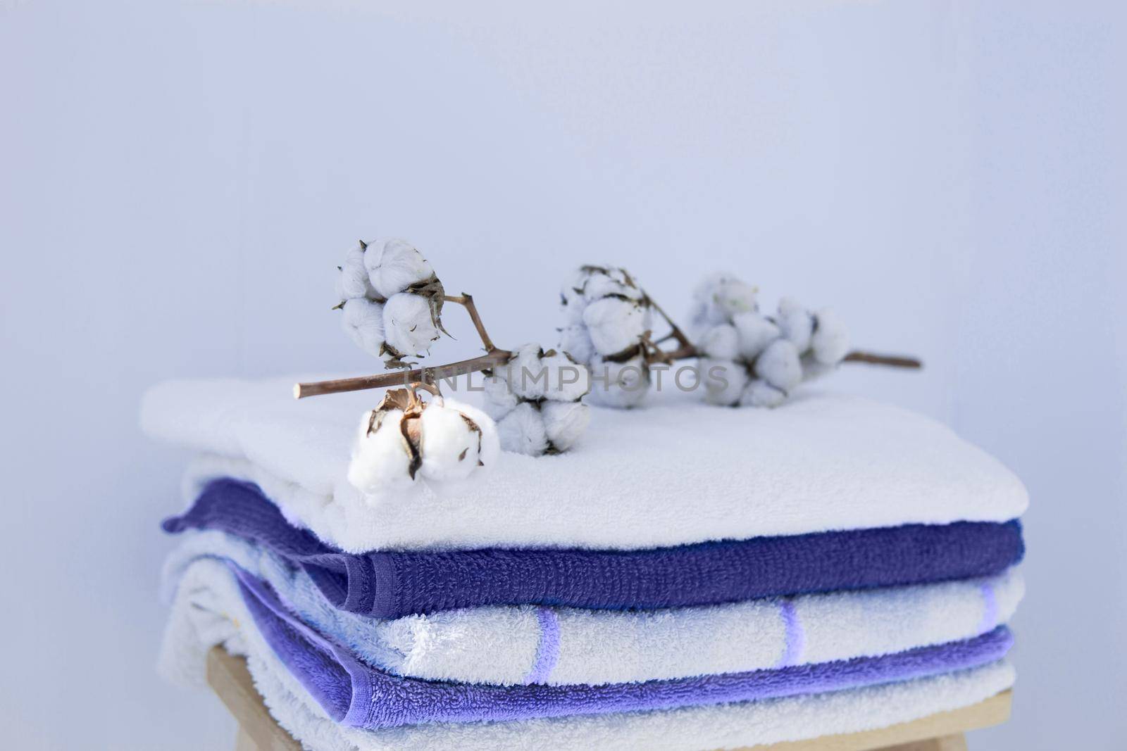 stack of clean bath towels colorful cotton Terry textiles with a sprig of cotton folded on a wooden chair against a white wall nap concept close-up country style. Demonstrating the colors of 2022 - Very Peri