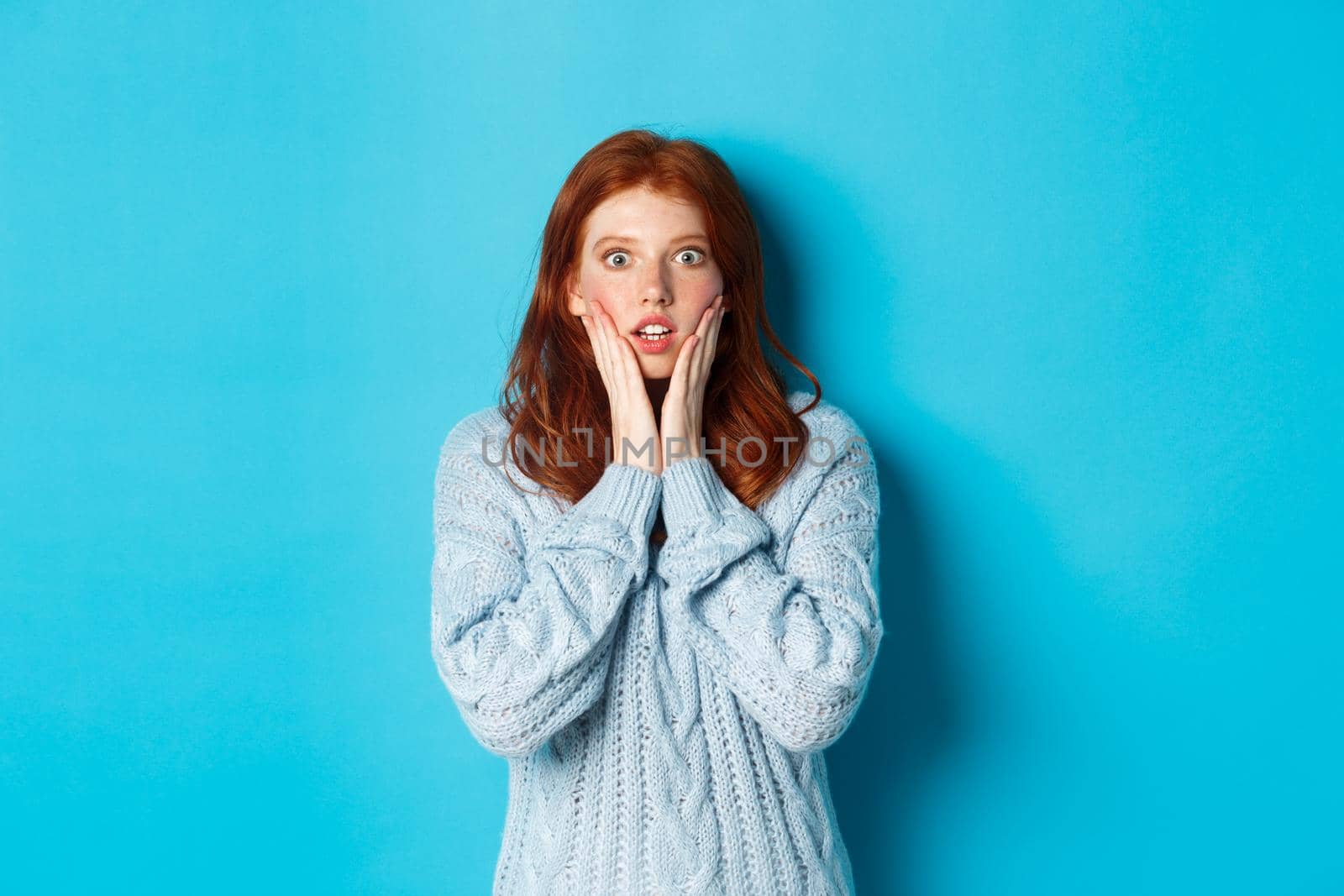 Shocked redhead girl staring at camera speechless, express disbelief and amazement, standing in sweater against blue background by Benzoix
