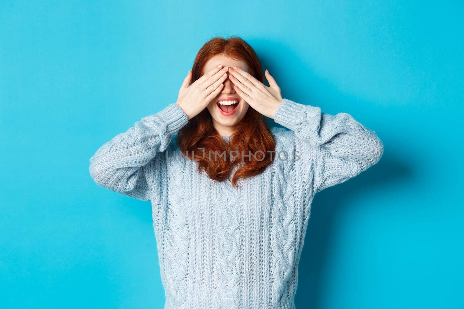 Cheerful redhead female model close eyes and waiting for christmas gift, holding hands on face and smiling amused, anticipating surprise, standing over blue background.