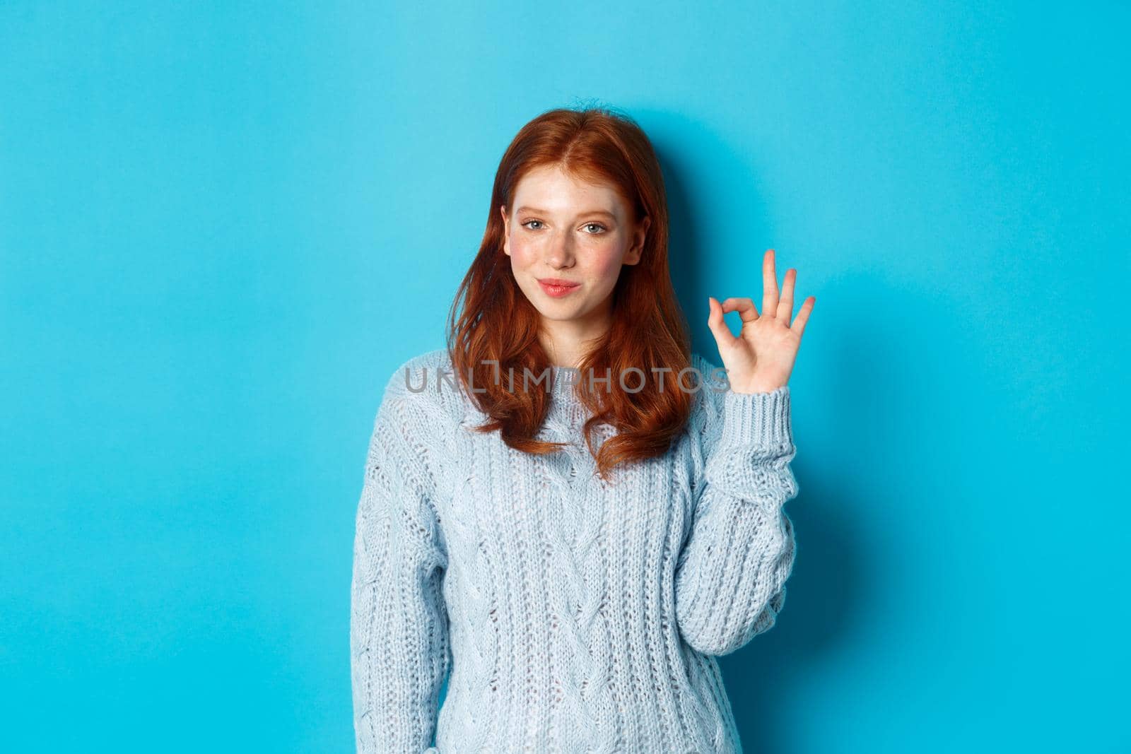 Confident redhead girl assuring you, showing okay sign and smiling, saying yes, approve and agree, standing over blue background by Benzoix