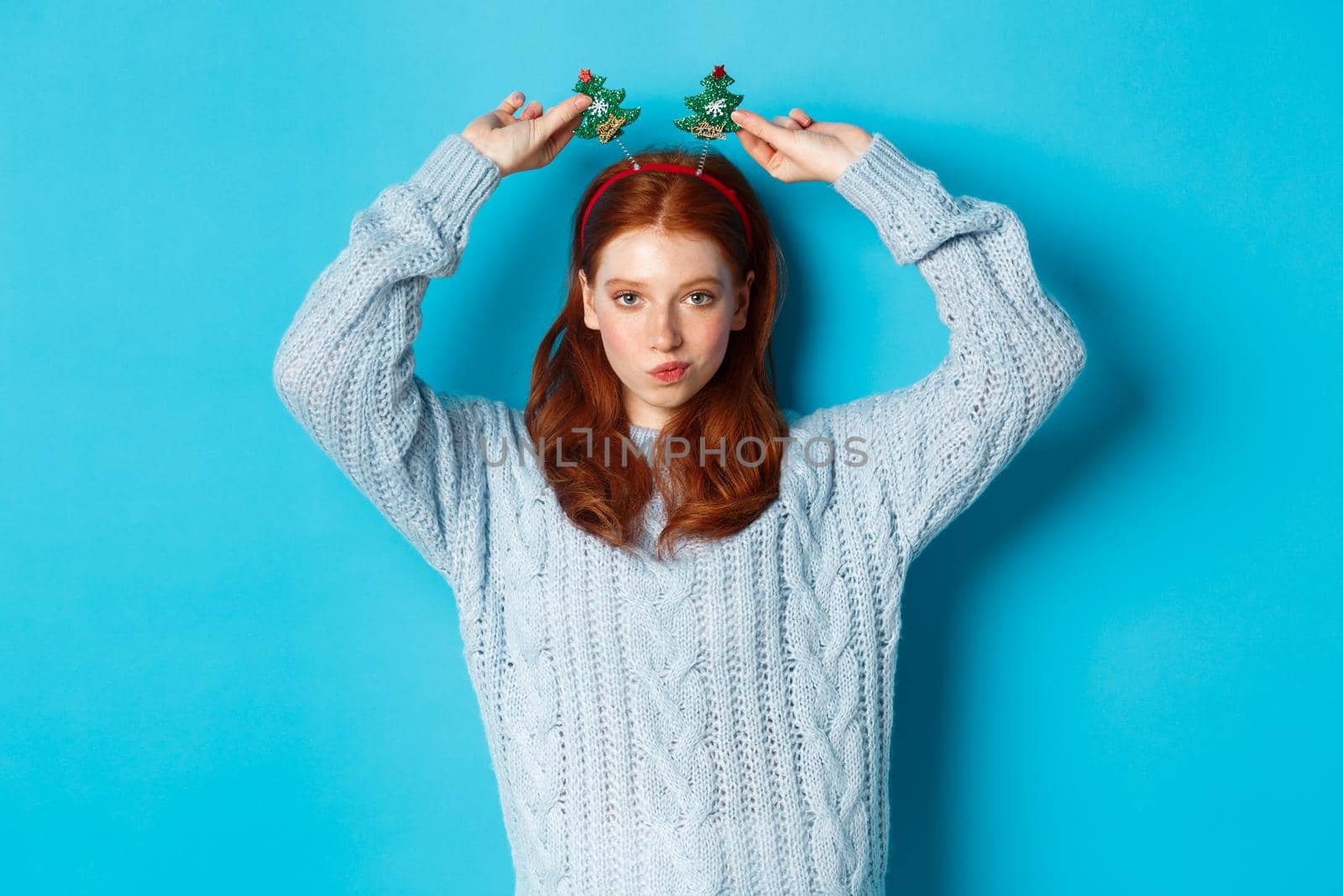 Winter holidays and Christmas sales concept. Beautiful redhead female model celebrating New Year, wearing funny party headband and sweater, smiling at camera.