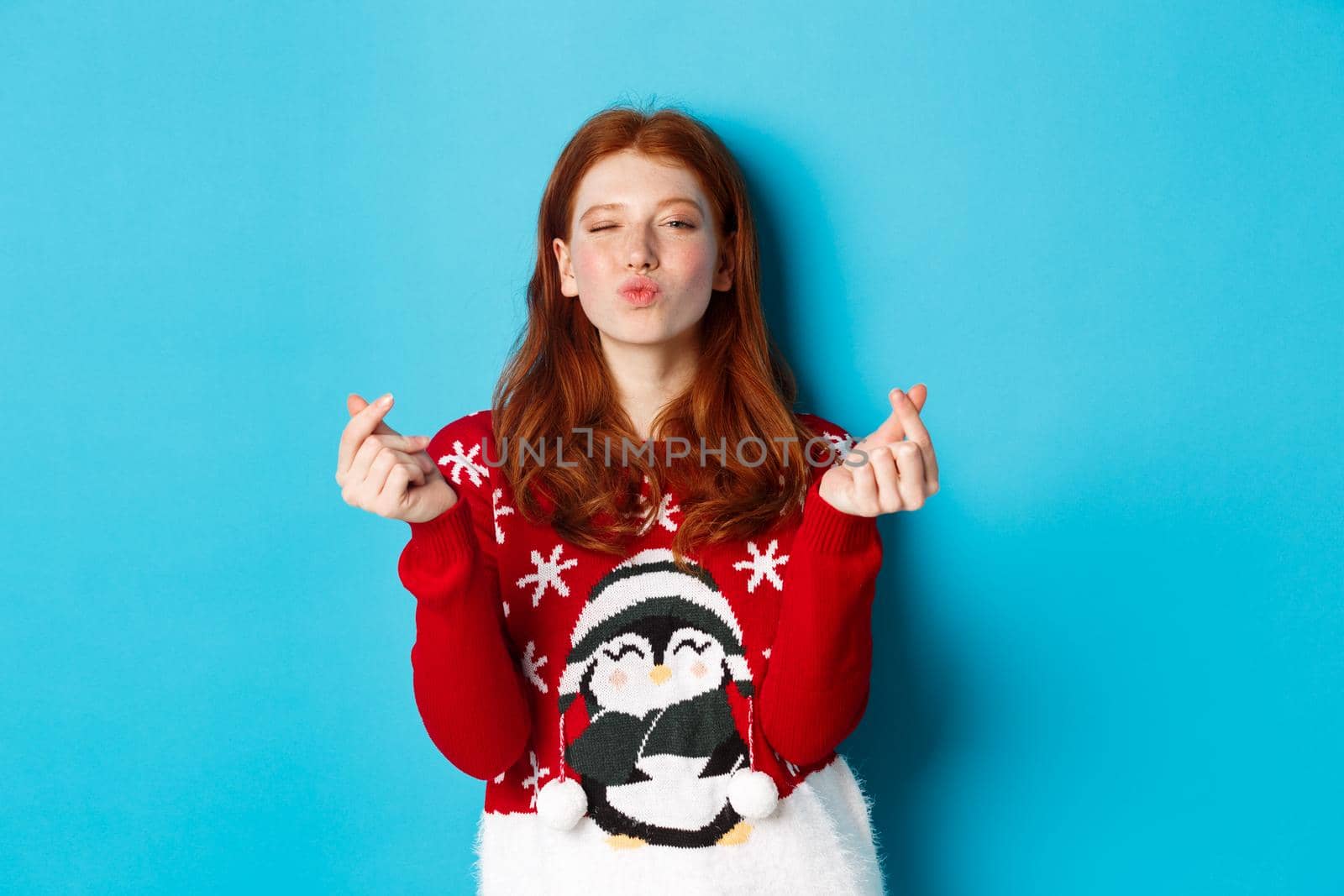 Winter holidays and Christmas Eve concept. Lovely redhead girl in xmas sweater, showing heart sign and pucker lips for kiss, standing over blue background. Copy space
