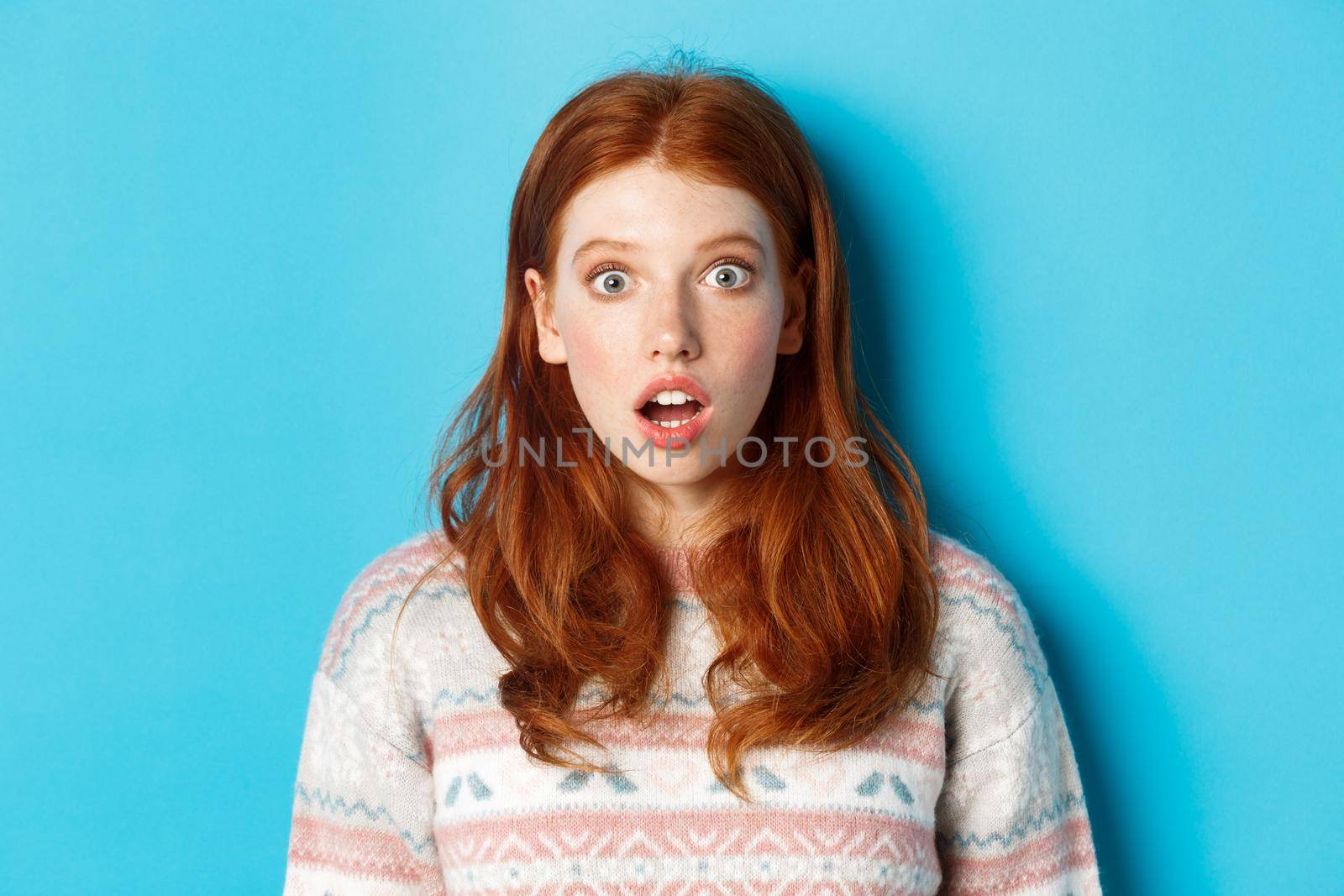 Close-up of shocked redhead girl drop jaw in awe, staring with amazement at camera, standing against blue background.