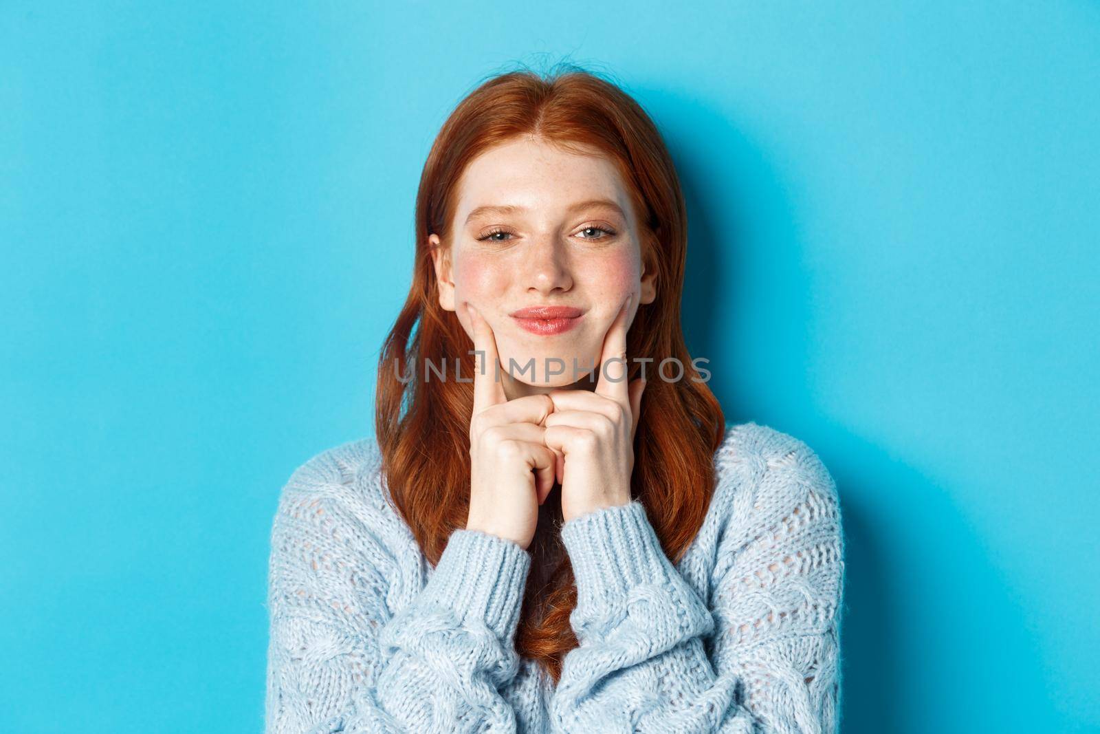 Close-up of cute and silly girl with red hair poking cheeks, showing dimples and smiling, standing against blue background by Benzoix