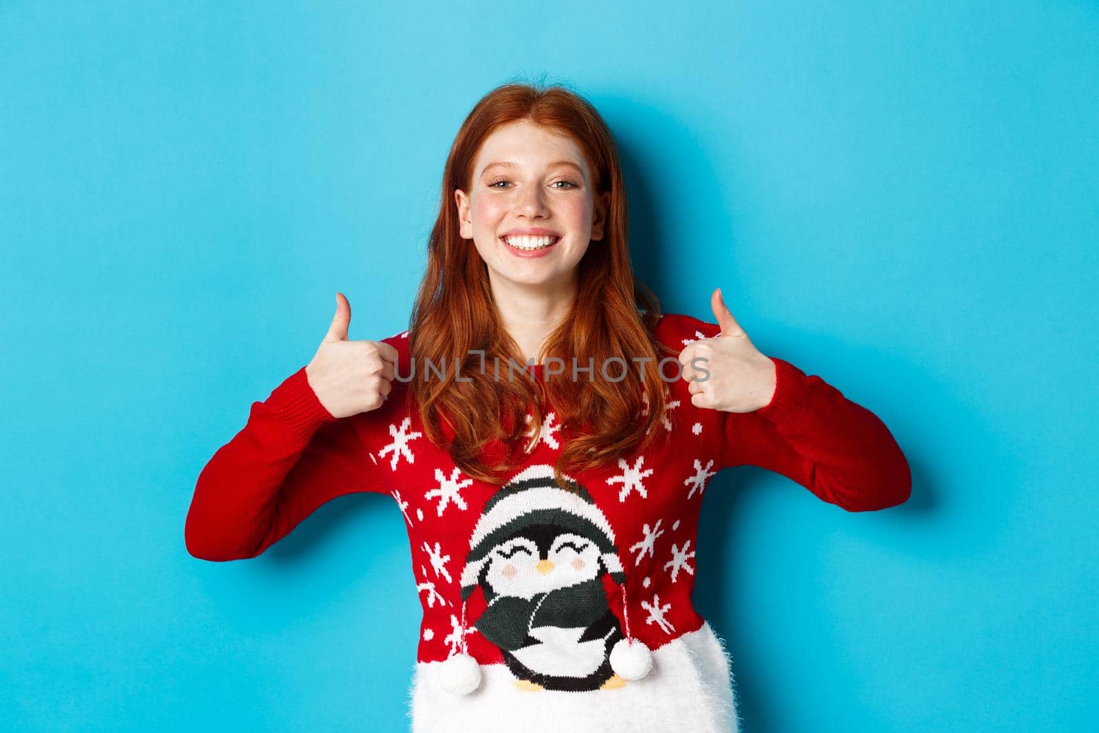 Winter holidays and Christmas Eve concept. Happy smiling girl with red hair, showing thumbs up in approval, praise good product, recommending, standing in xmas sweater.