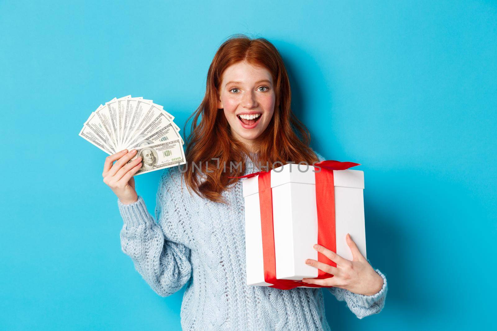 Christmas and shopping concept. Happy redhead woman holding money and big xmas present, showing dollars and gift, smiling pleased, standing over blue background.