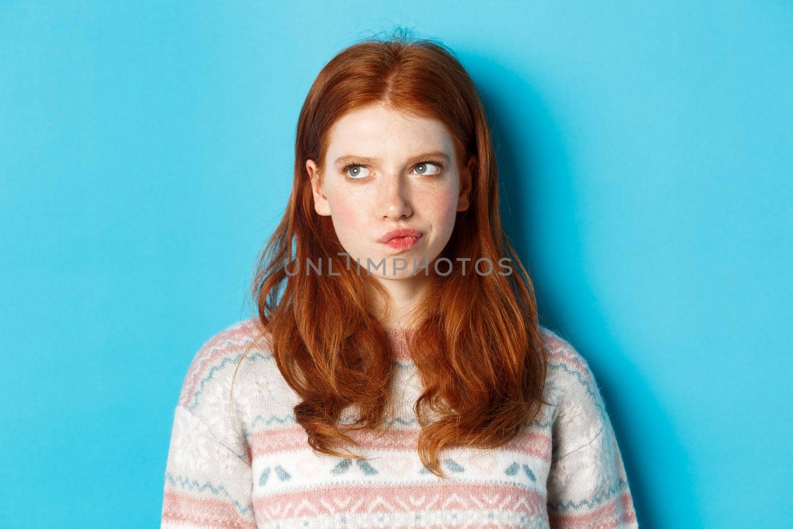 Serious redhead girl thinking, grimacing and frowning while pondering, looking left at copy space, standing over blue background by Benzoix