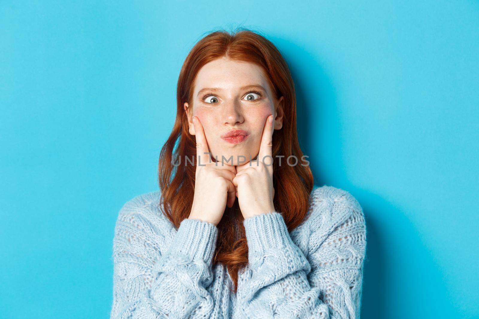 Close-up of funny redhead teen girl making faces, squinting and pocking cheeks, standing against blue background.