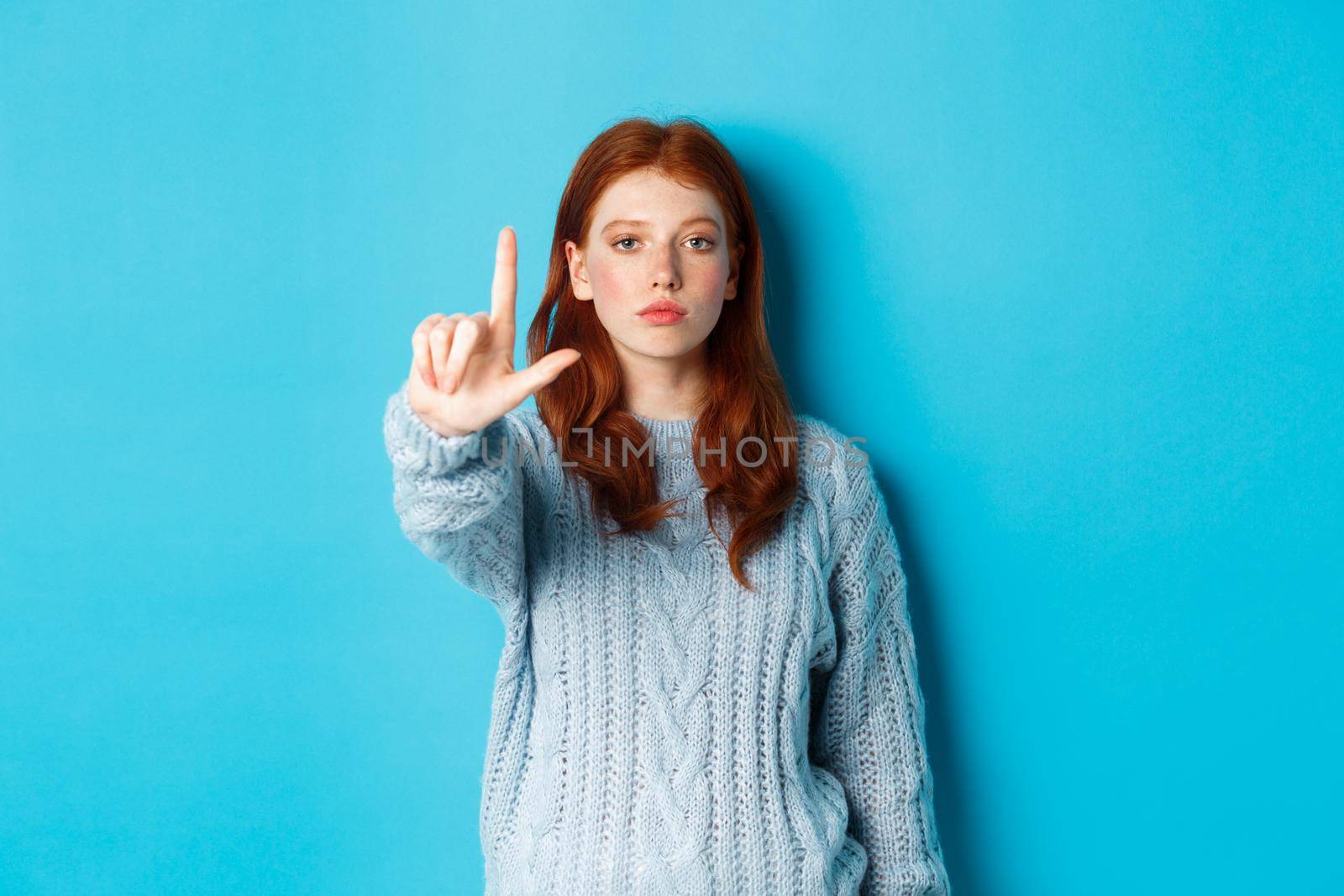 Serious redhead girl in sweater showing taboo gesture, extending one finger, shaking forefinger to disapprove, disagree and forbid something, blue background.