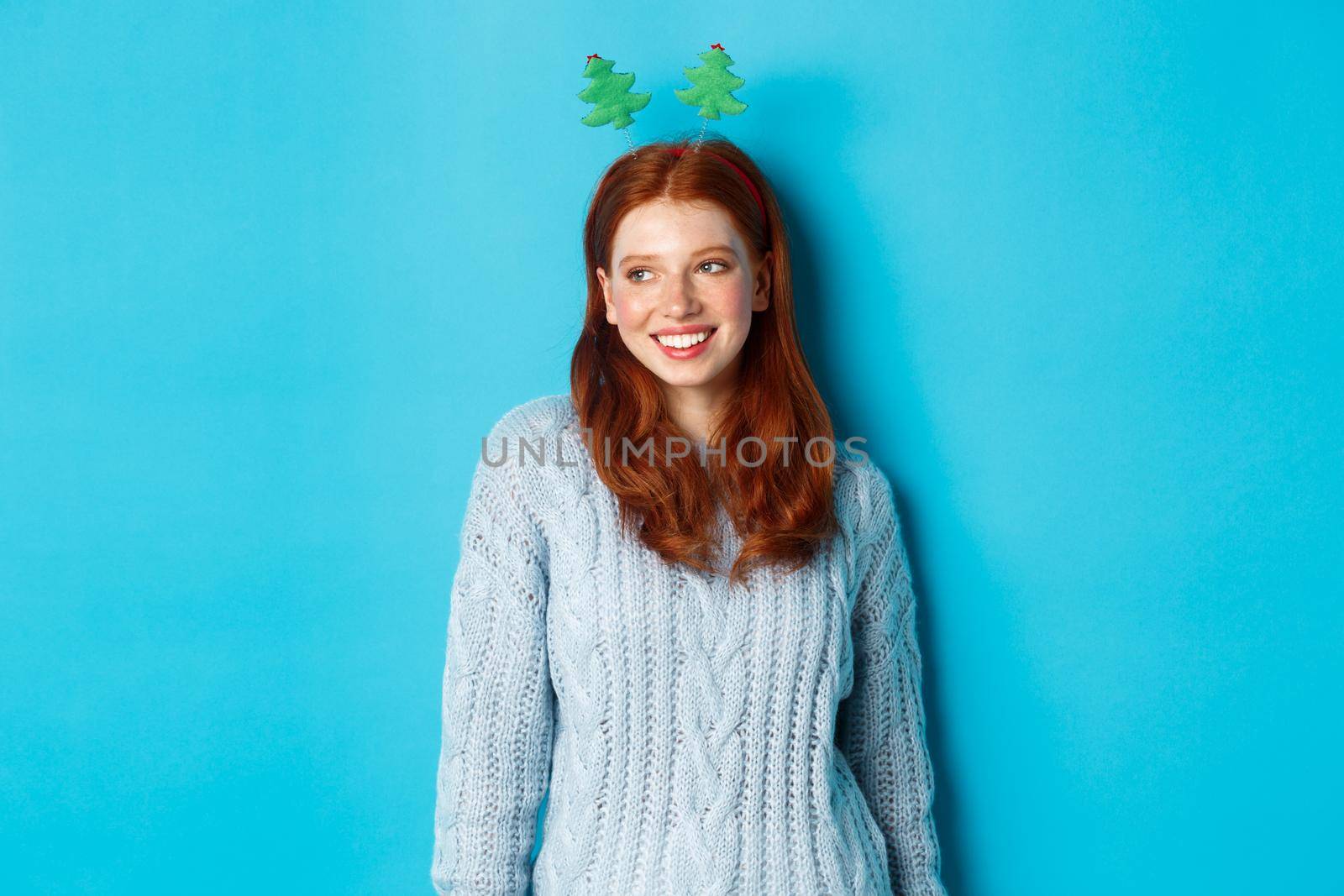 Winter holidays and Christmas sales concept. Cute redhead girl in funny New Year headband smiling, looking left at logo, standing over blue background.