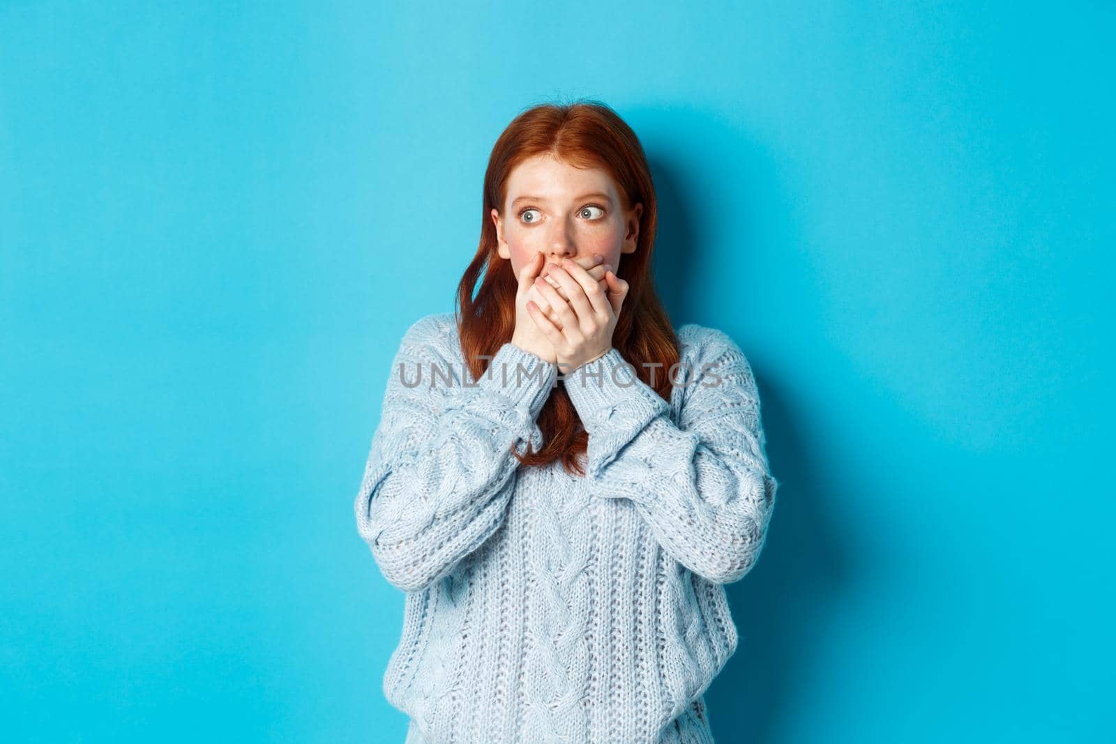 Shocked and anxious redhead girl staring left scared, covering mouth and gasping, standing over blue background in sweater.