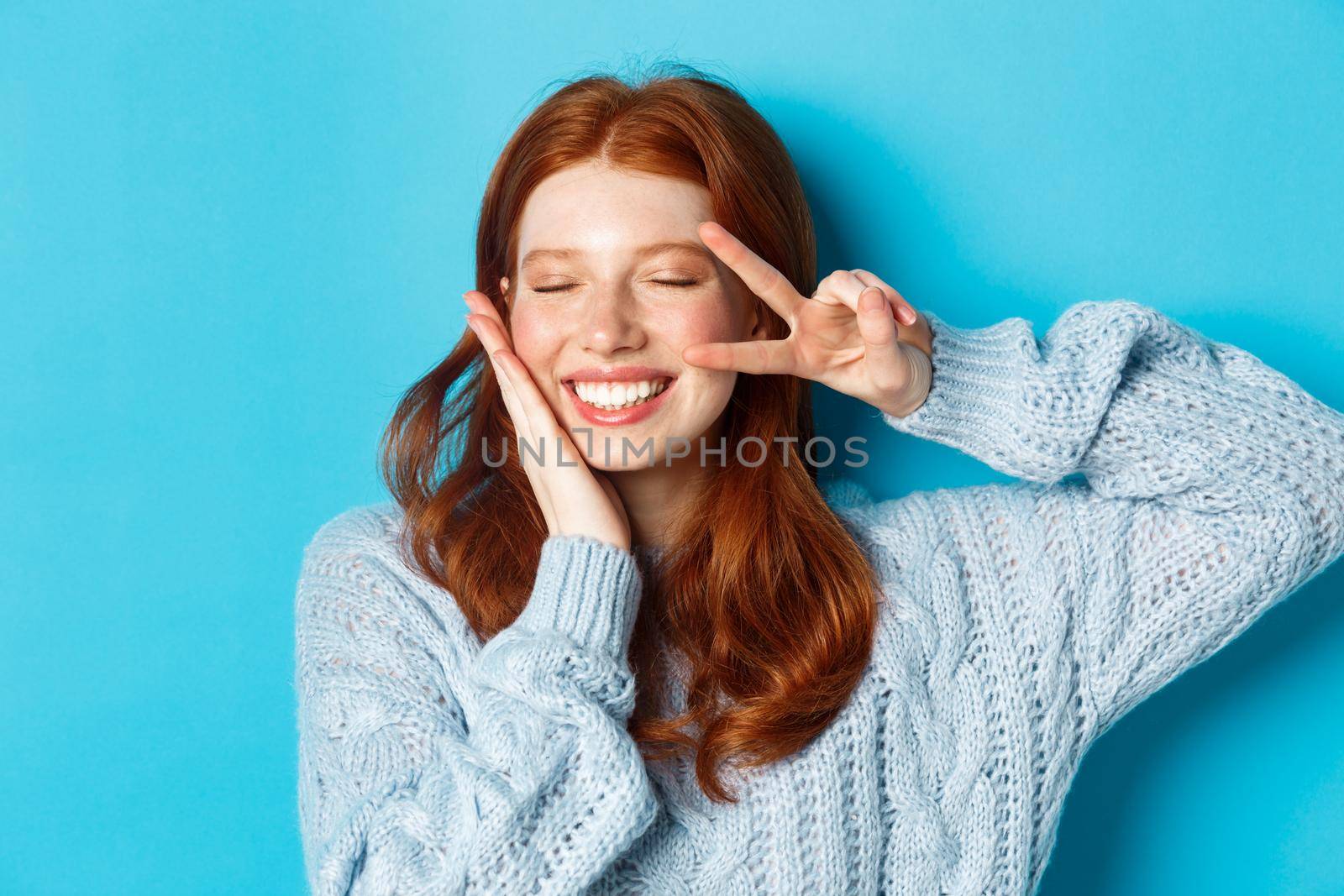Close-up of beautiful smiling girl with red hair, showing peace kawaii sign, standing with eyes closed over blue background.