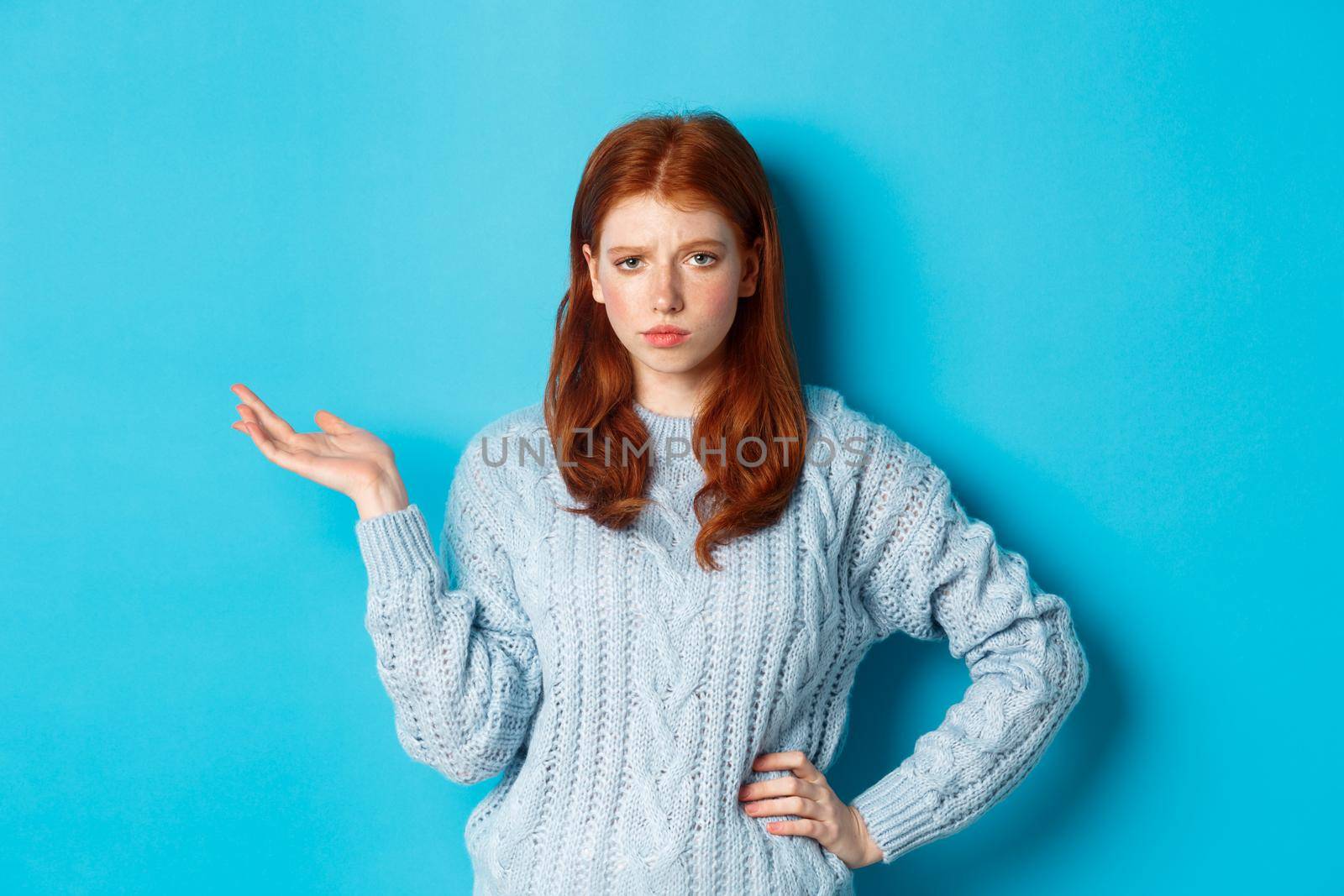 Skeptical teenage girl looking unamused, raising hand in so what gesture, staring at something with careless face, standing over blue background.