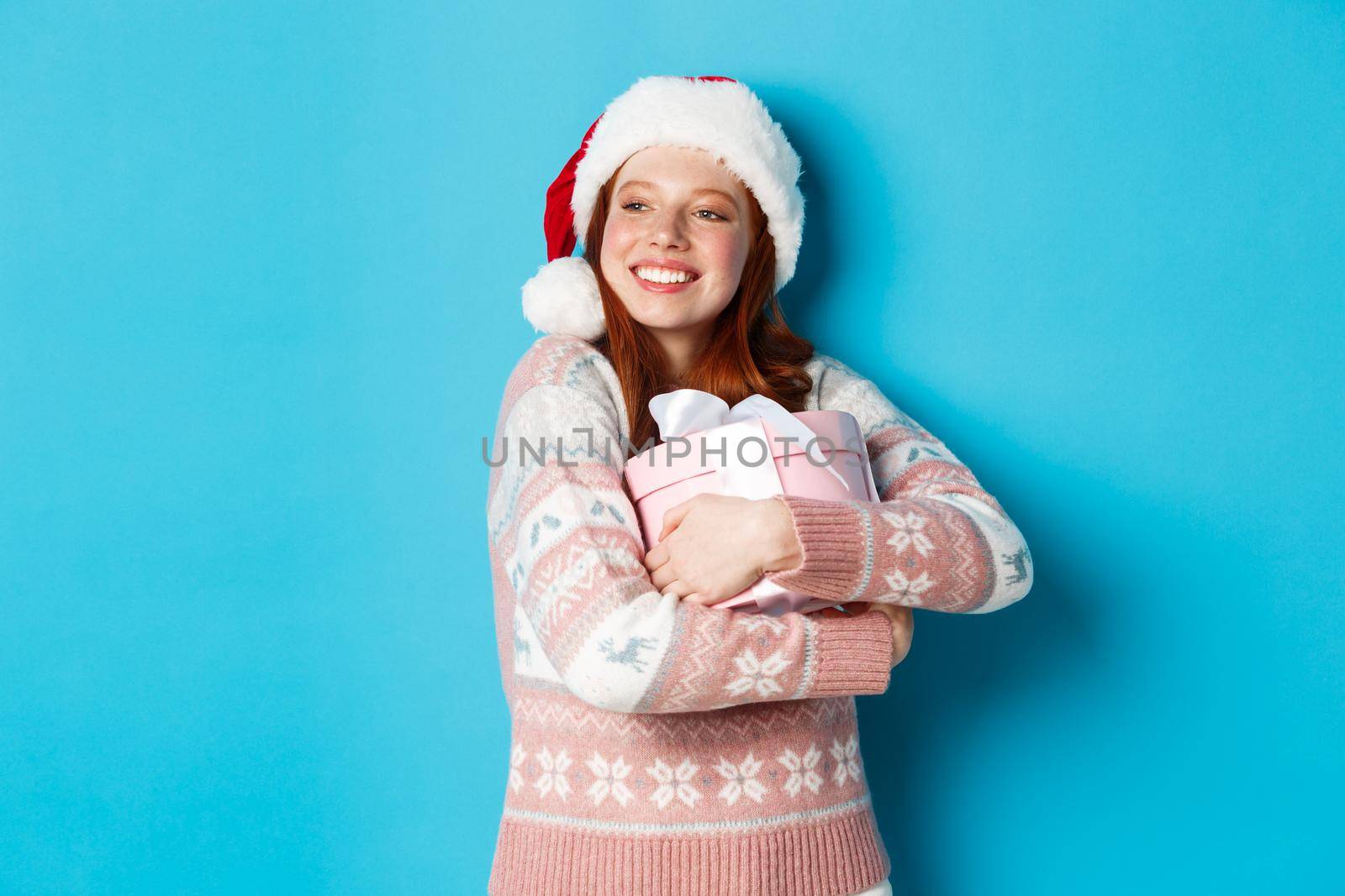 Winter and celebration concept. Dreamy redhead girl in santa hat hugging her christmas gift and looking left, smiling happy, standing over blue background by Benzoix