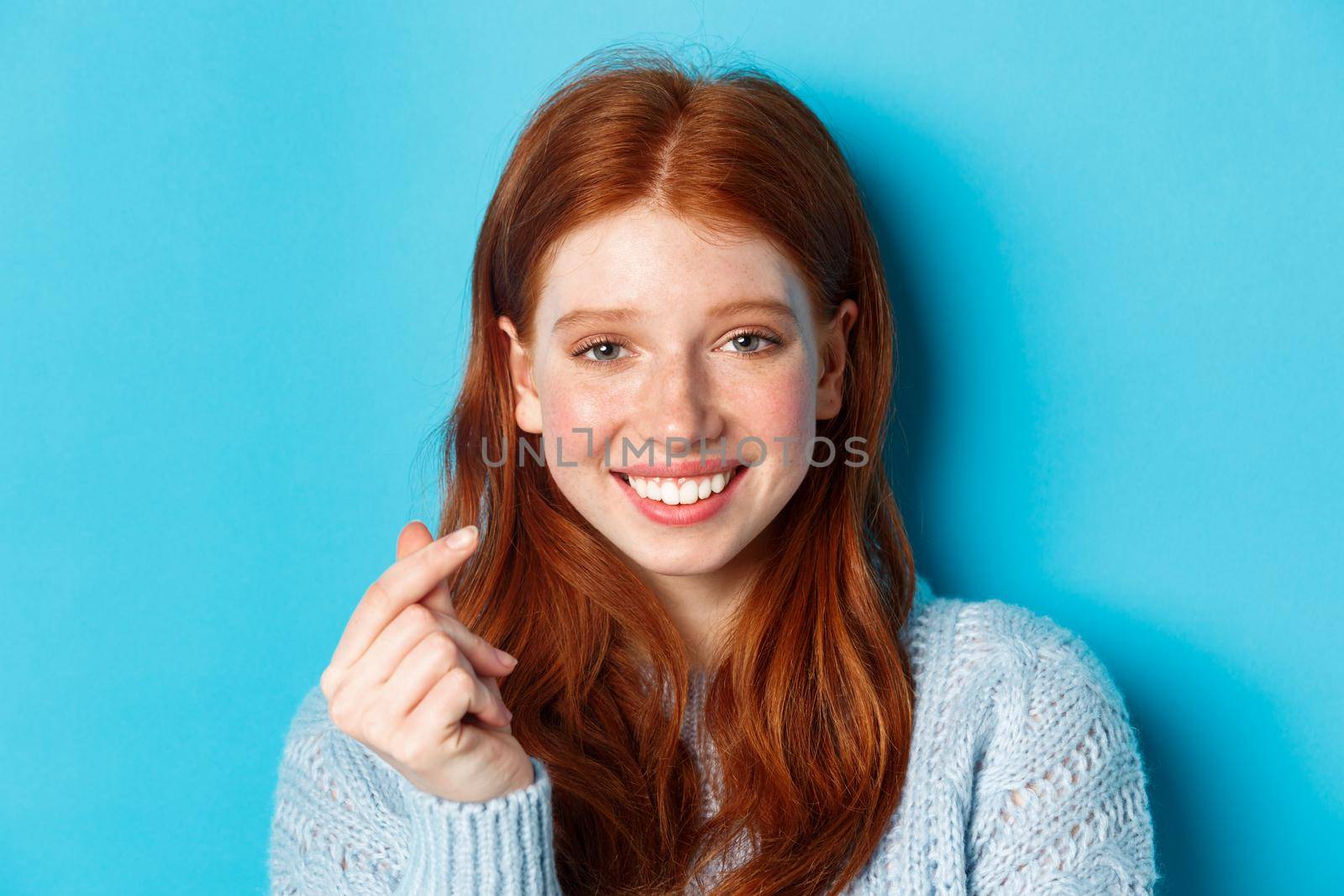 Headshot of cute caucasian woman with red hair and freckles showing heart sign and smiling, standing against blue background by Benzoix