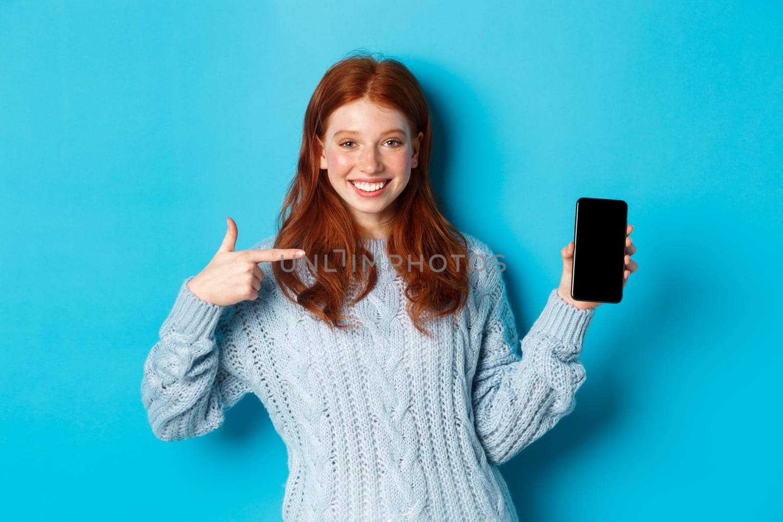 Satisfied redhead girl pointing at phone screen, showing smartphone app or online promo and smiling, standing in sweater against blue background.