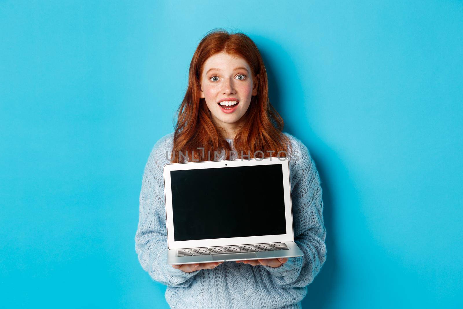 Excited redhead female freelancer showing laptop screen, staring at camera amazed, standing with computer against blue background.