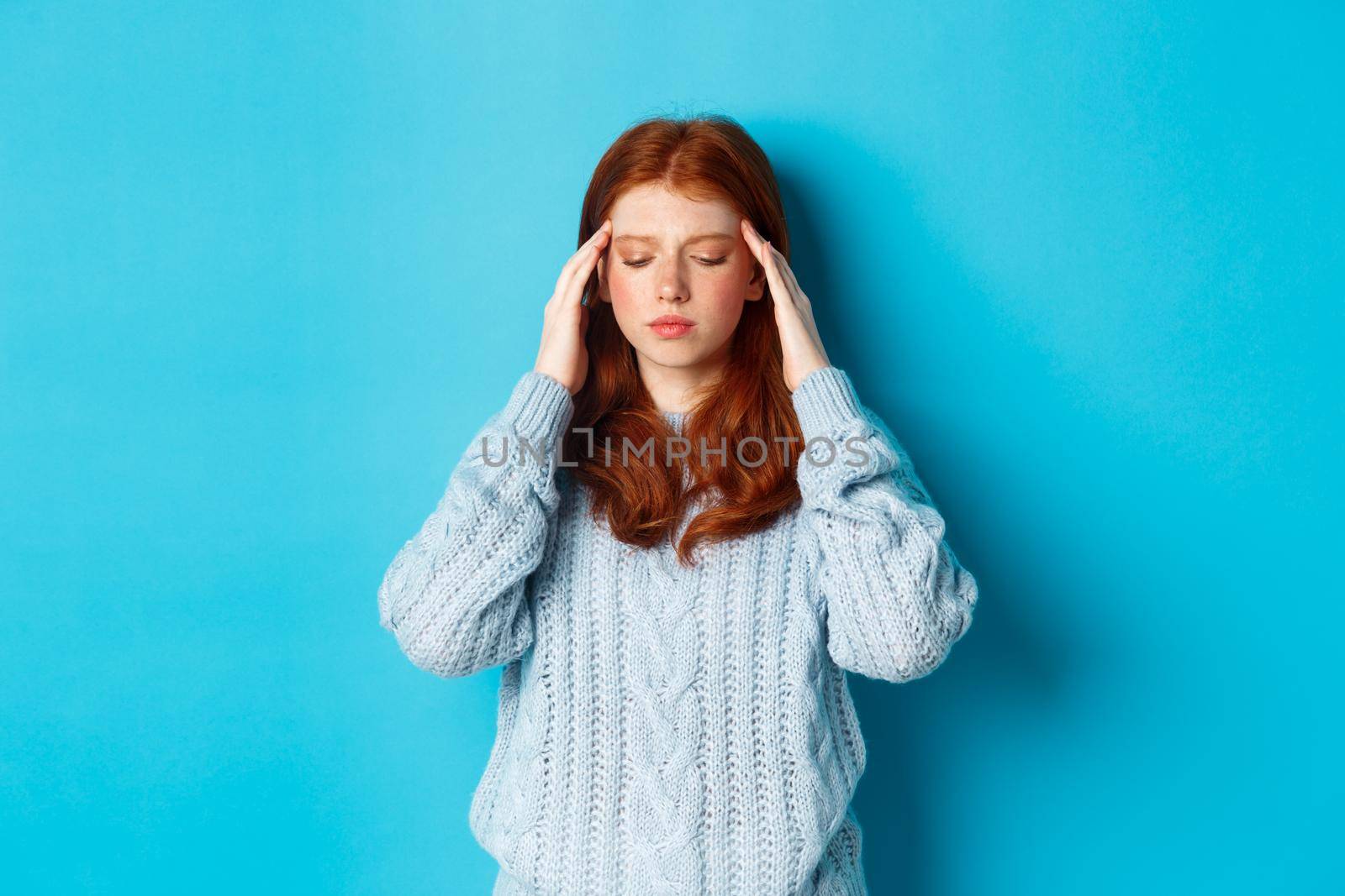 Distressed teenage redhead girl touching head, looking down with troubled face expression, standing against blue background, have problem.