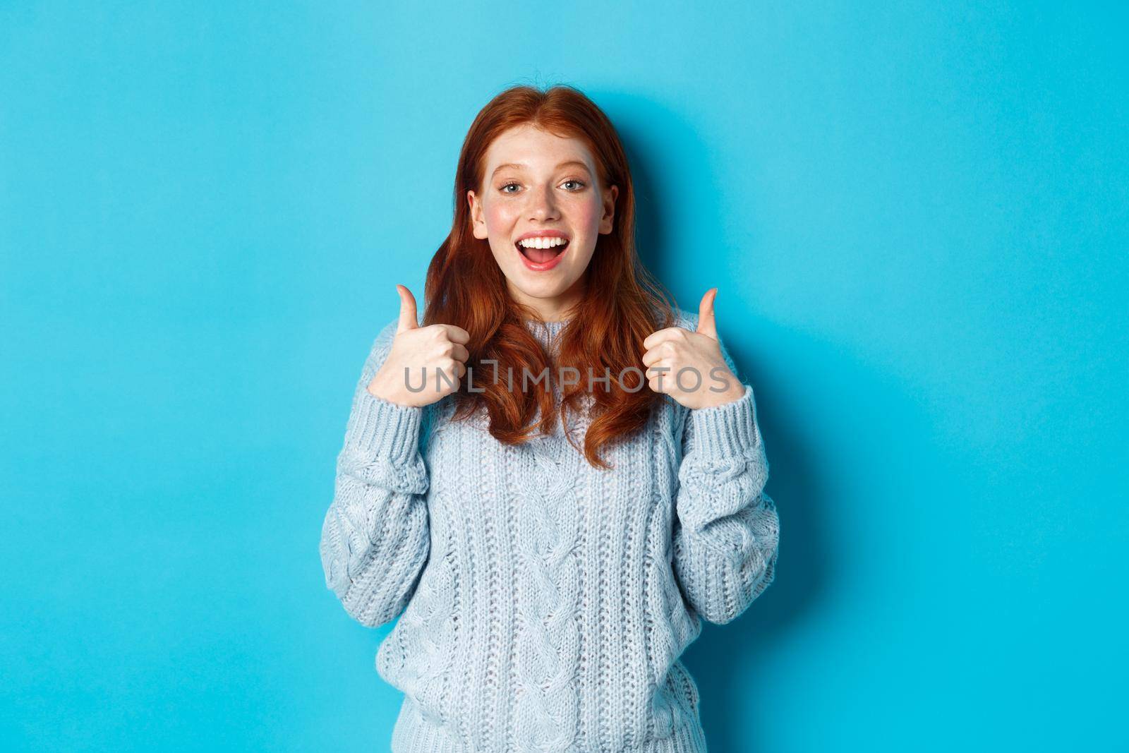 Supportive redhead girl in sweater, showing thumbs-up and looking amazed, praising good choice, standing over blue background.