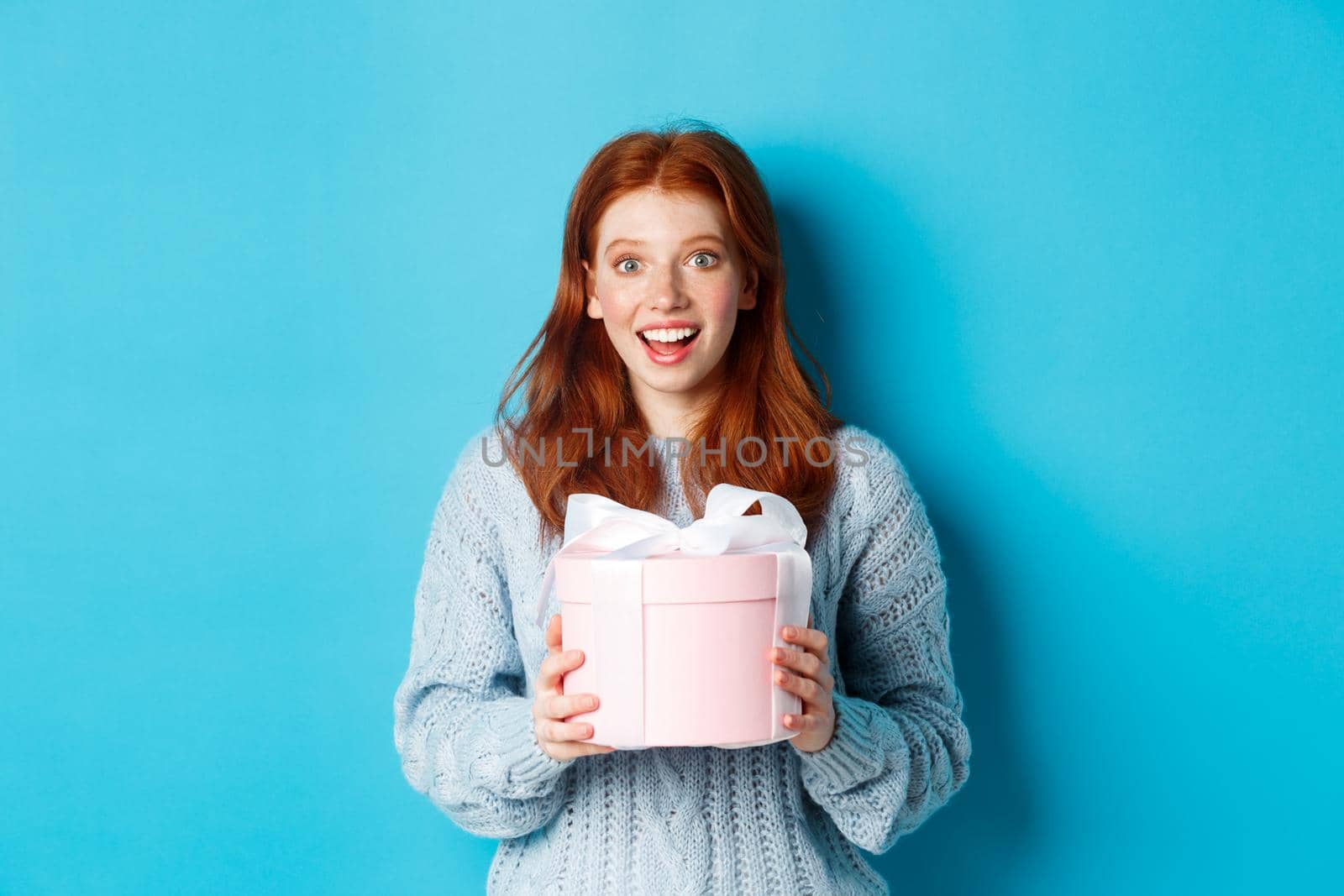 Surprised redhead girl receiving valentines gift, holding box with present and staring at camera amazed, wearing sweater, standing over blue background.