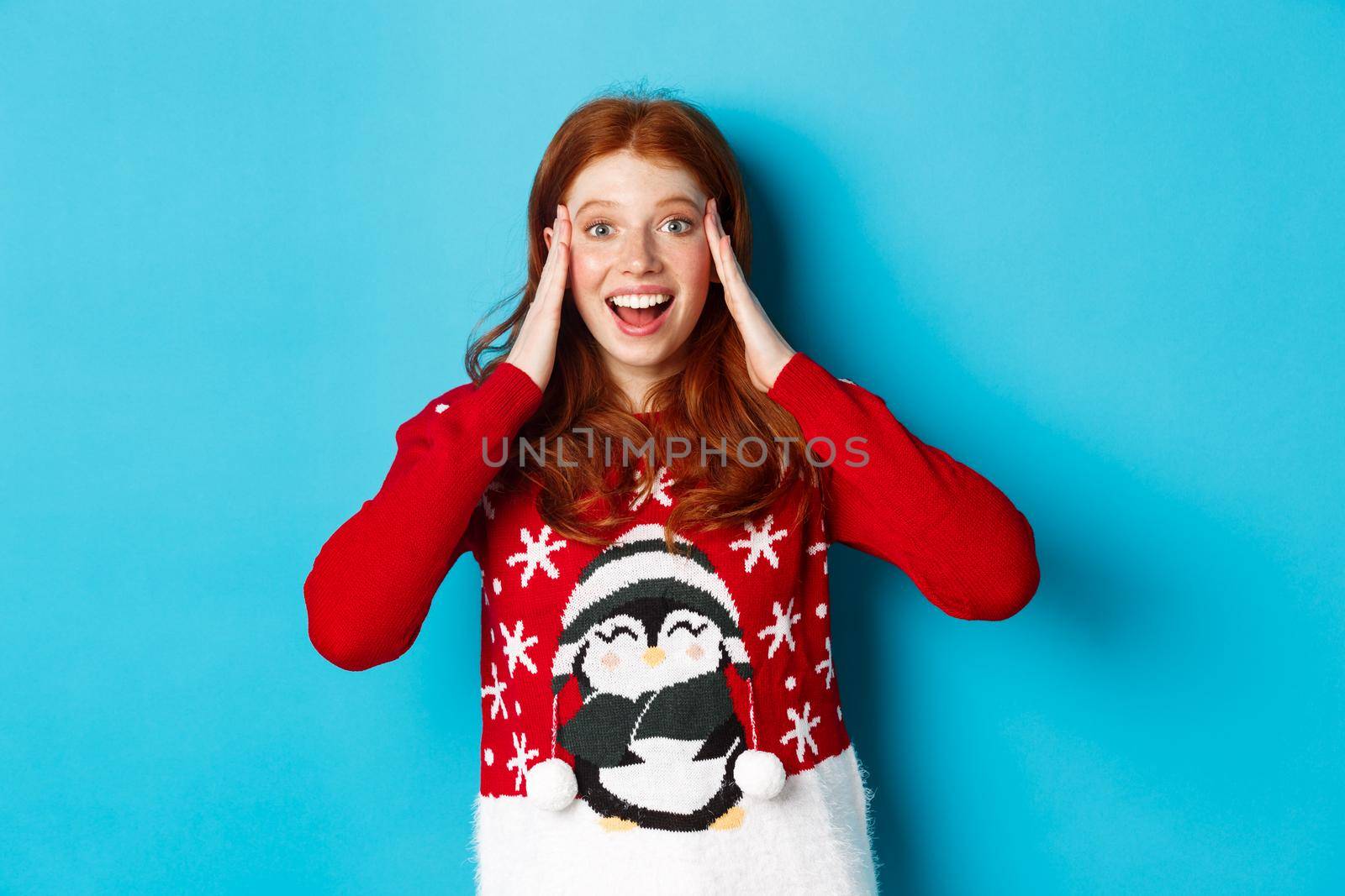 Winter holidays and Christmas Eve concept. Surprised and excited redhead girl, holding hands on head and stare with disbelief at xmas gift, standing over blue background by Benzoix
