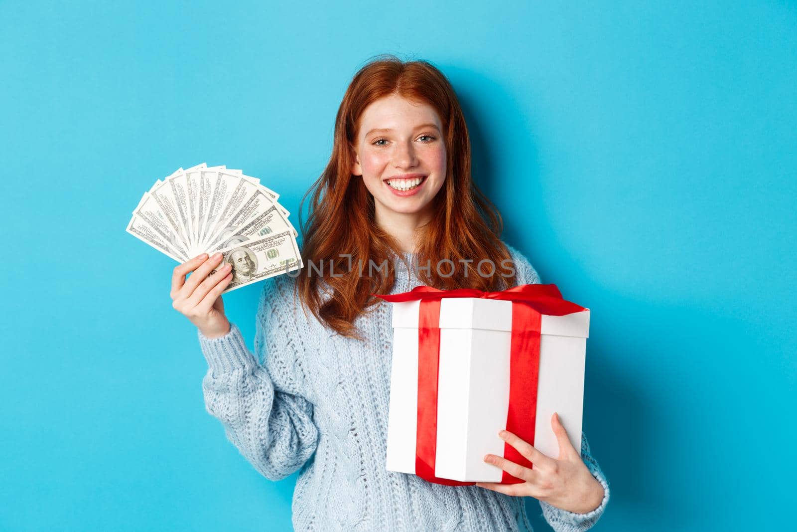 Young redhead woman holding Christmas gift box and money, smiling pleased, standing over blue background.