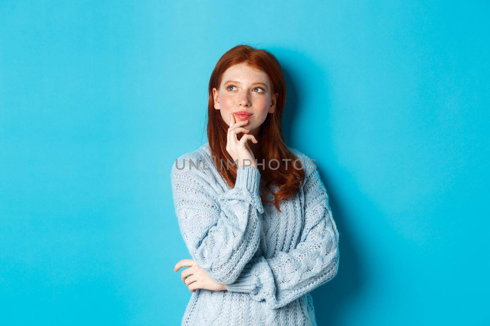 Thoughtful cute woman with red hair, looking upper left corner logo and thinking, imaging something, standing over blue background by Benzoix