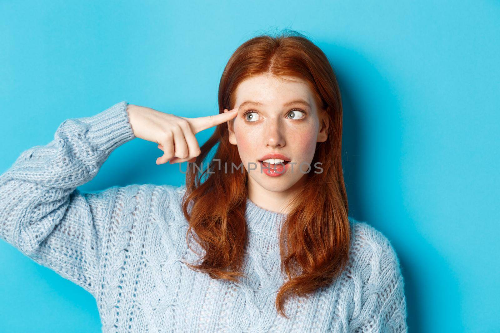 Close-up of cute redhead girl searching for solution, pointing at head and looking left at logo, standing over blue background.