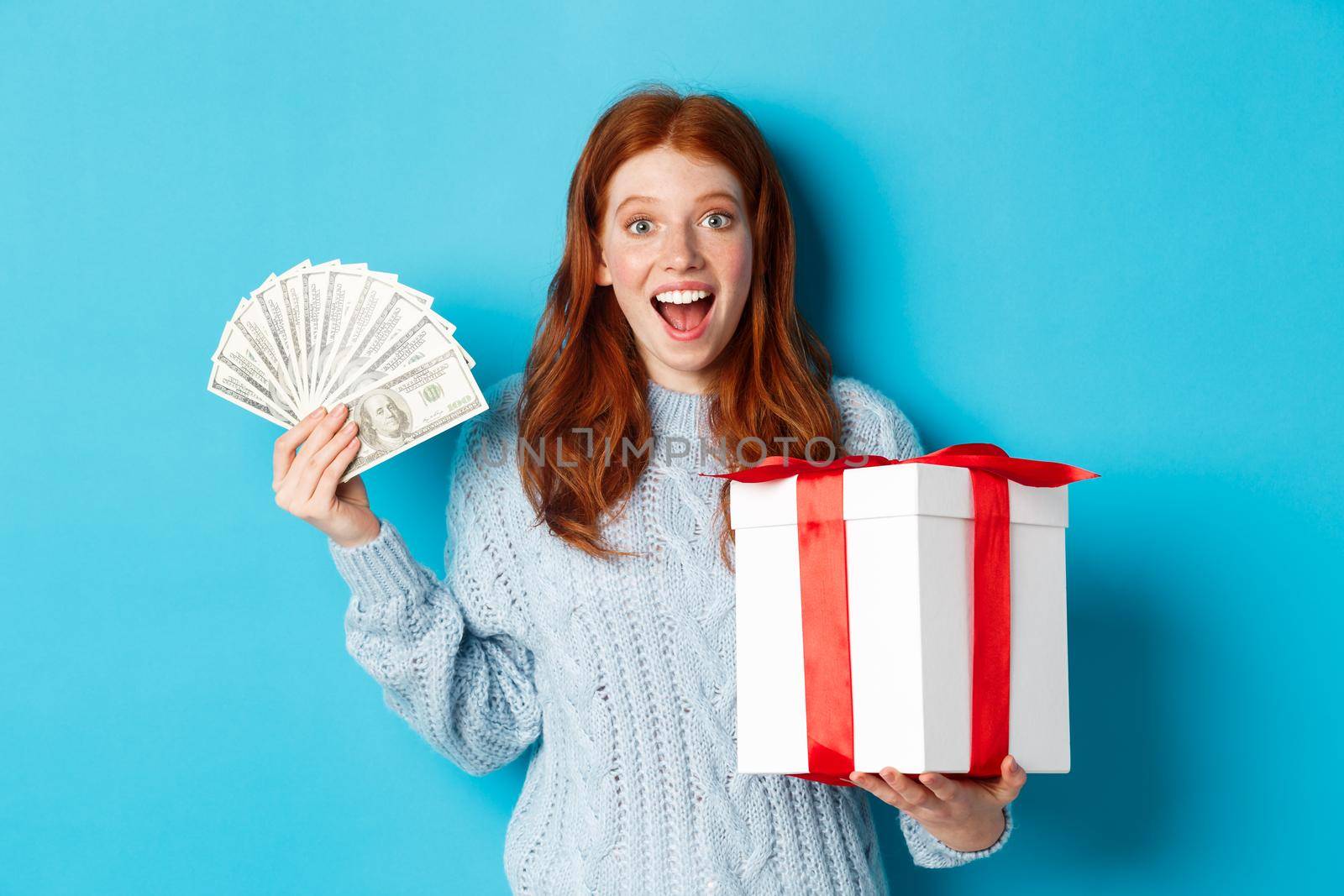 Christmas and shopping concept. Happy redhead woman holding money and big xmas present, showing dollars and gift, smiling pleased, standing over blue background by Benzoix