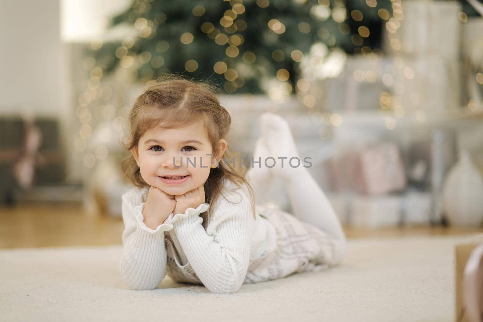 Adorable little girl lying on her front nearby fir tree and smile. Christmas mood at home with lot of presents.