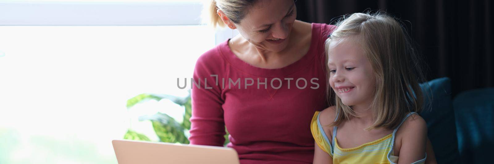 Smiling mom and daughter sitting on couch and looking at laptop. Joint viewing of educational programs for children in family concept