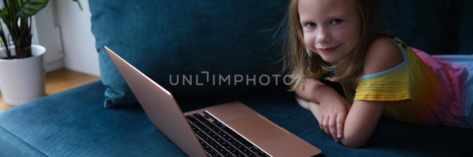 Little smiling girl lies on couch with laptop. Baby apps digital technology concept