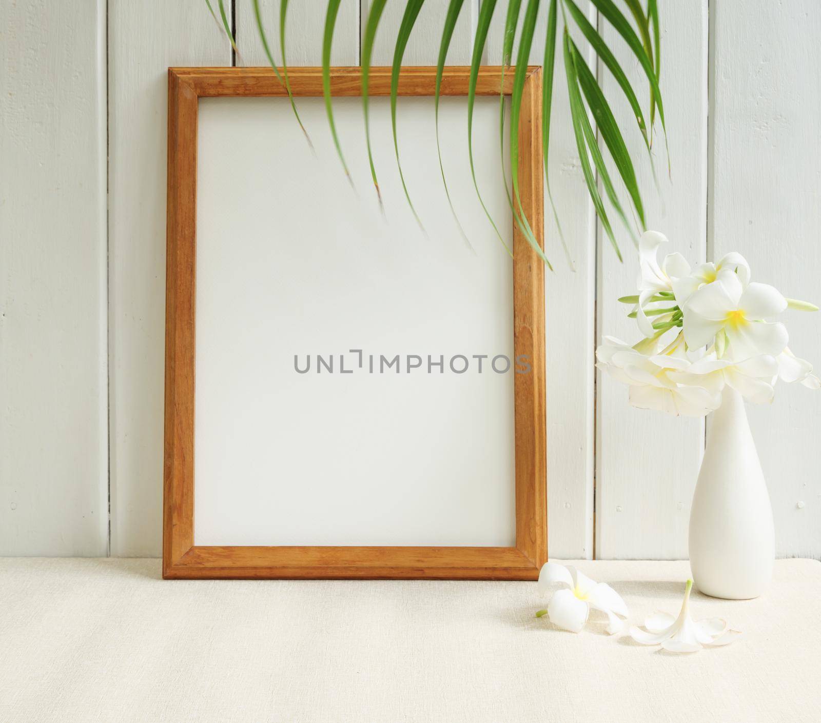 Mock up poster wooden frame and beautiful Frangipani tropical flower in modern white vase on beige table and wood wall background with  green palm leaf