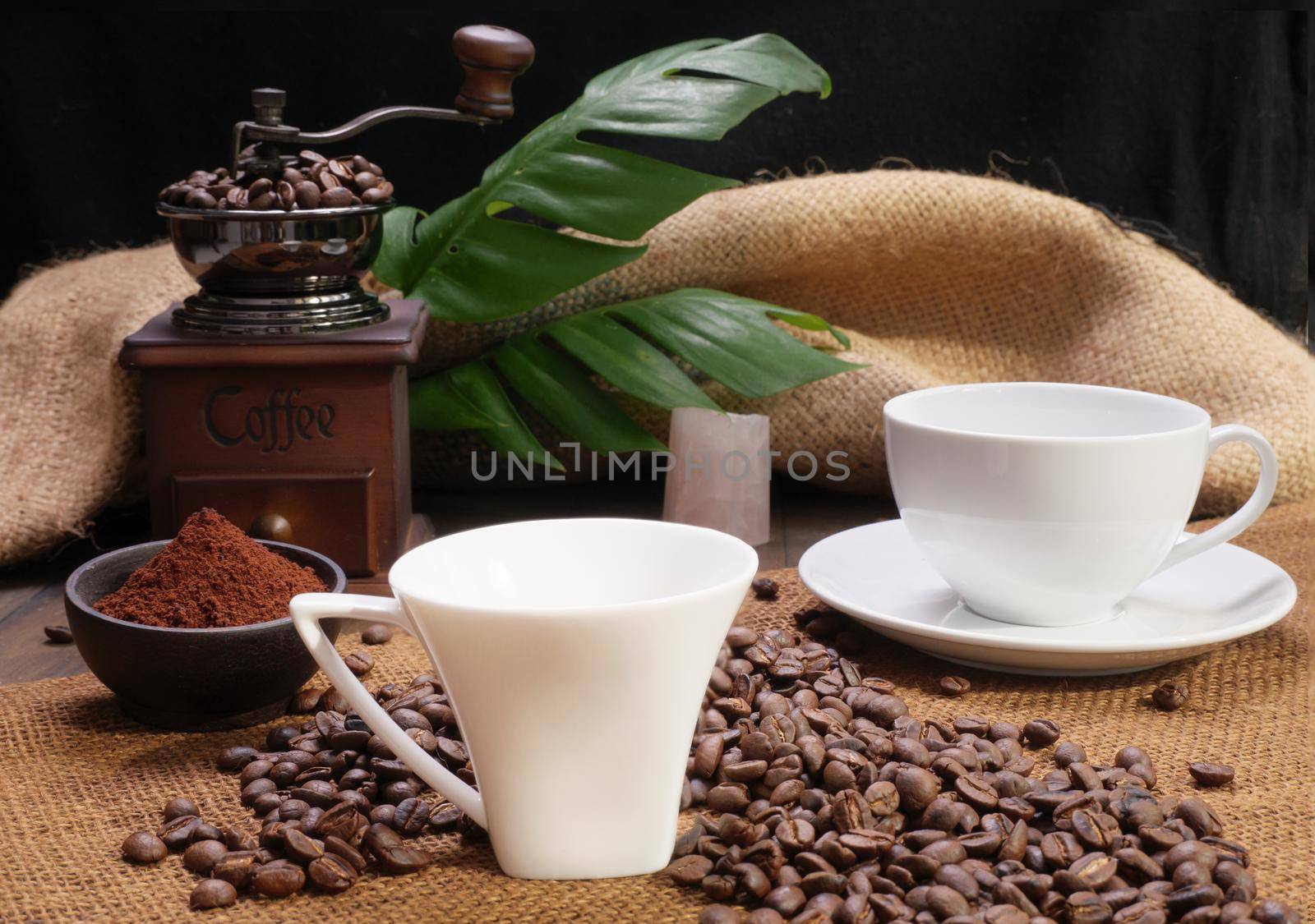 Two coffee cups,ground coffee ,roasted coffee beans,grinder, and monstera leave on wooden table with burlap  background