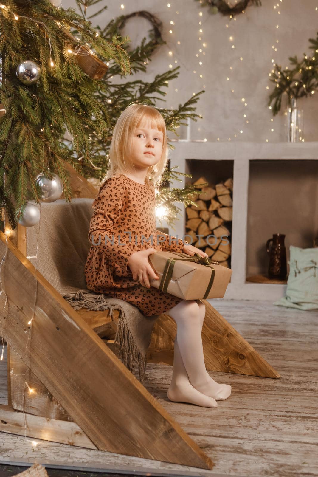 A little blonde girl is sitting on a wooden staircase in a Scandinavian interior decorated in a New Year's style. A child holds a Christmas gift in a craft package.