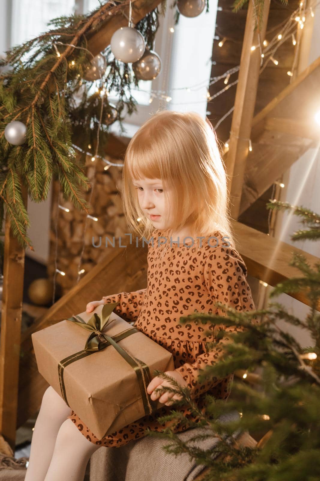 A little blonde girl is sitting on a wooden staircase in a Scandinavian interior decorated in a New Year's style. A child holds a Christmas gift in a craft package.