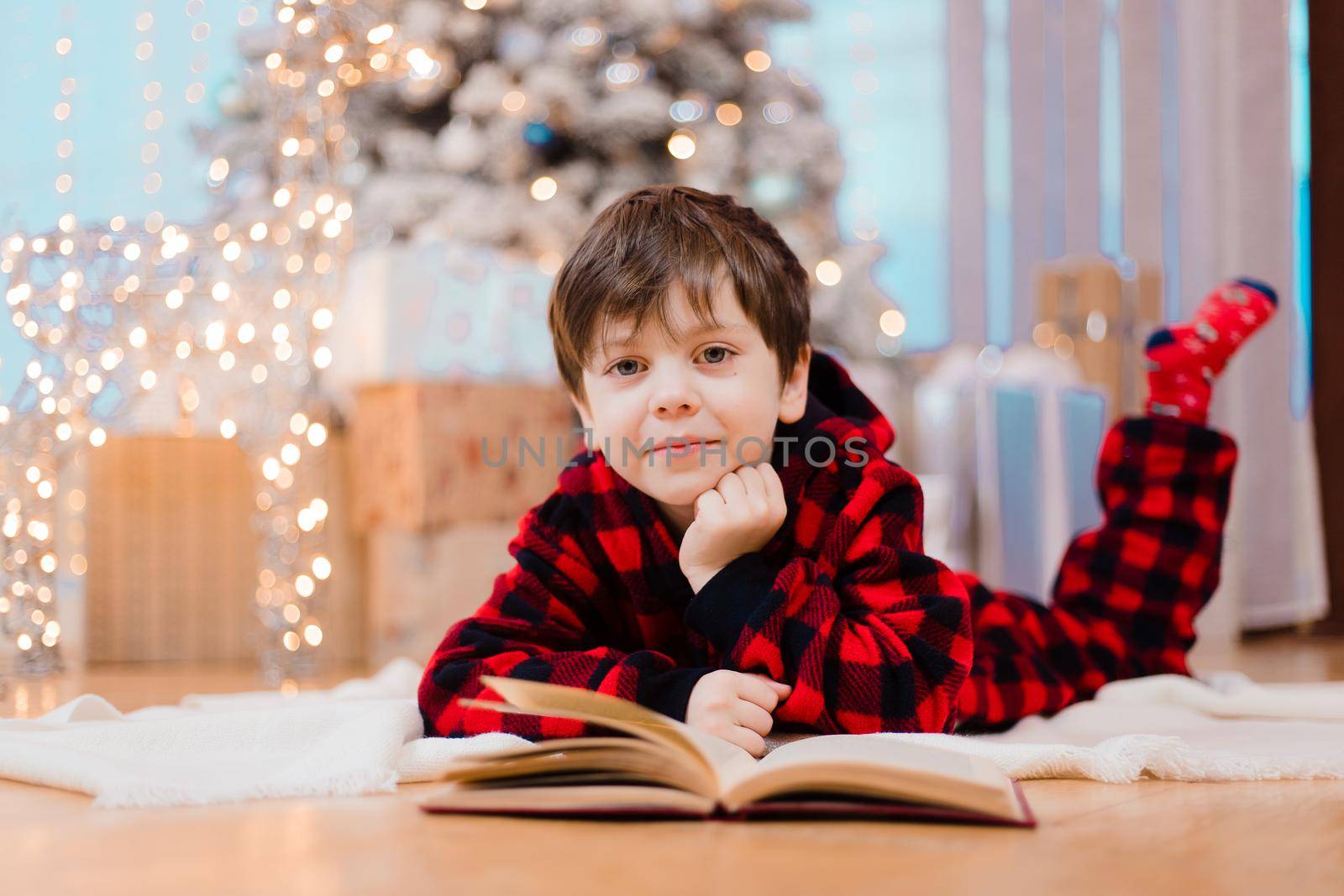 A boy in pajamas reads a book under a Christmas tree . New Year's mood. Reading books . Children's books. A gift for the new year