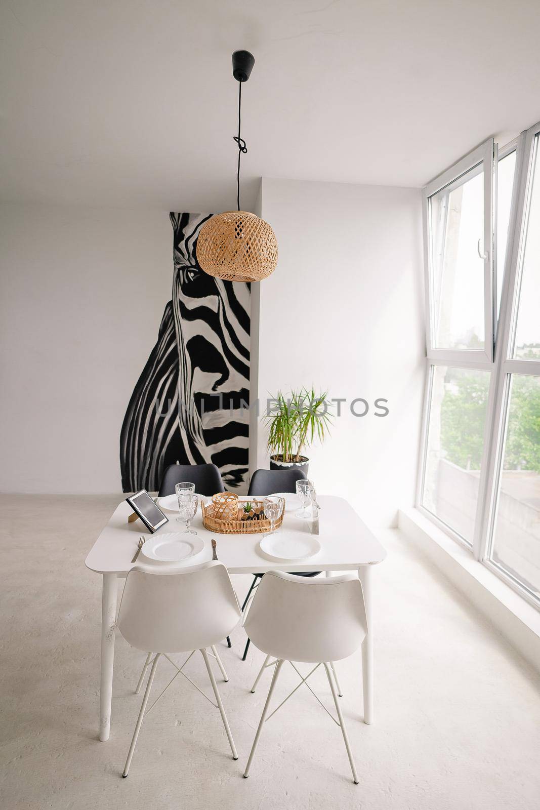 Interior Design of a Light Dining Room in a Minimalist Style with a Square Wooden Server White Table, Black and White Chairs, with Wicker Decorations on the Chandelier and on the Table, with a Large Window and a Huge Zebra Picture on the Wall. High quality photo