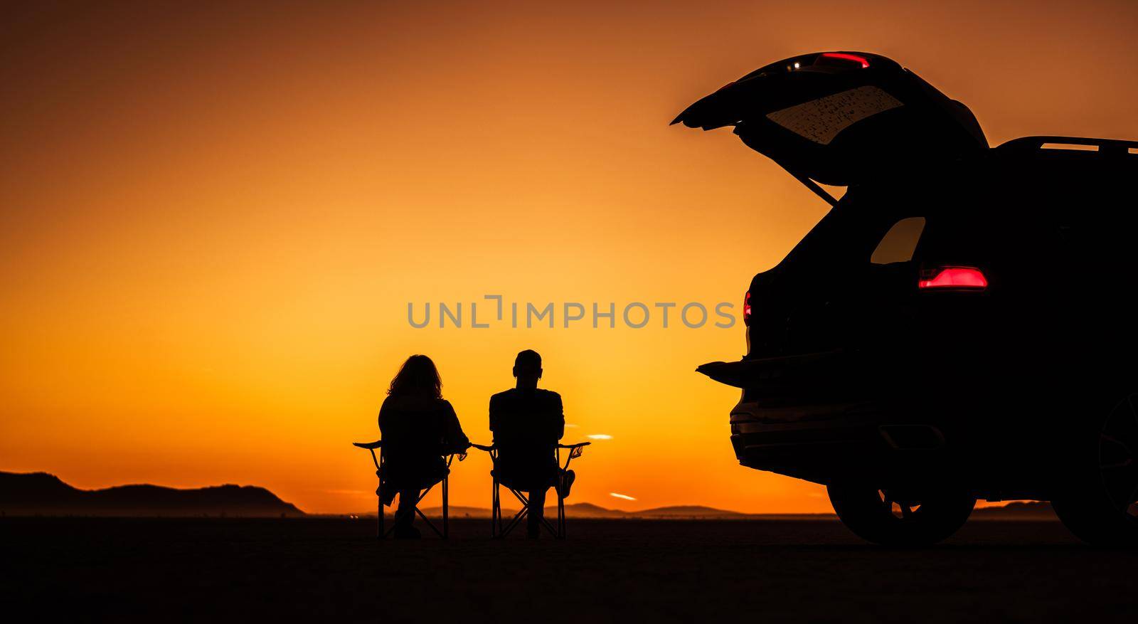 Couple Enjoying Their Life Watching Scenic Sunset on the Middle of a Desert Seating on Camping Chairs Next to Their Sport Utility Vehicle. Southern California USA