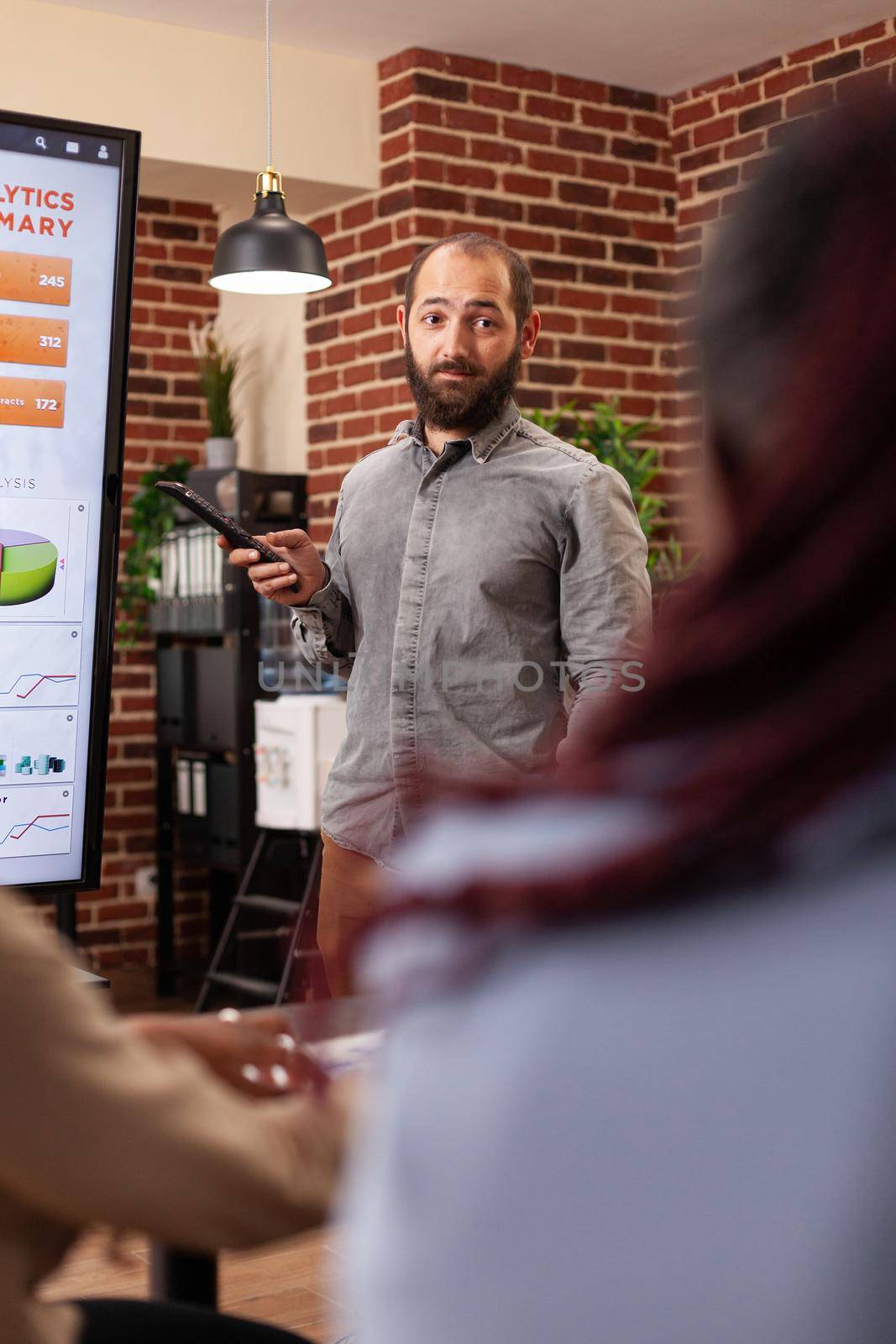 Entrepreneur man pointing at monitor explaining management statistics to businessteam working at company presentation in startup office. Diverse businesspeople planning business presentation